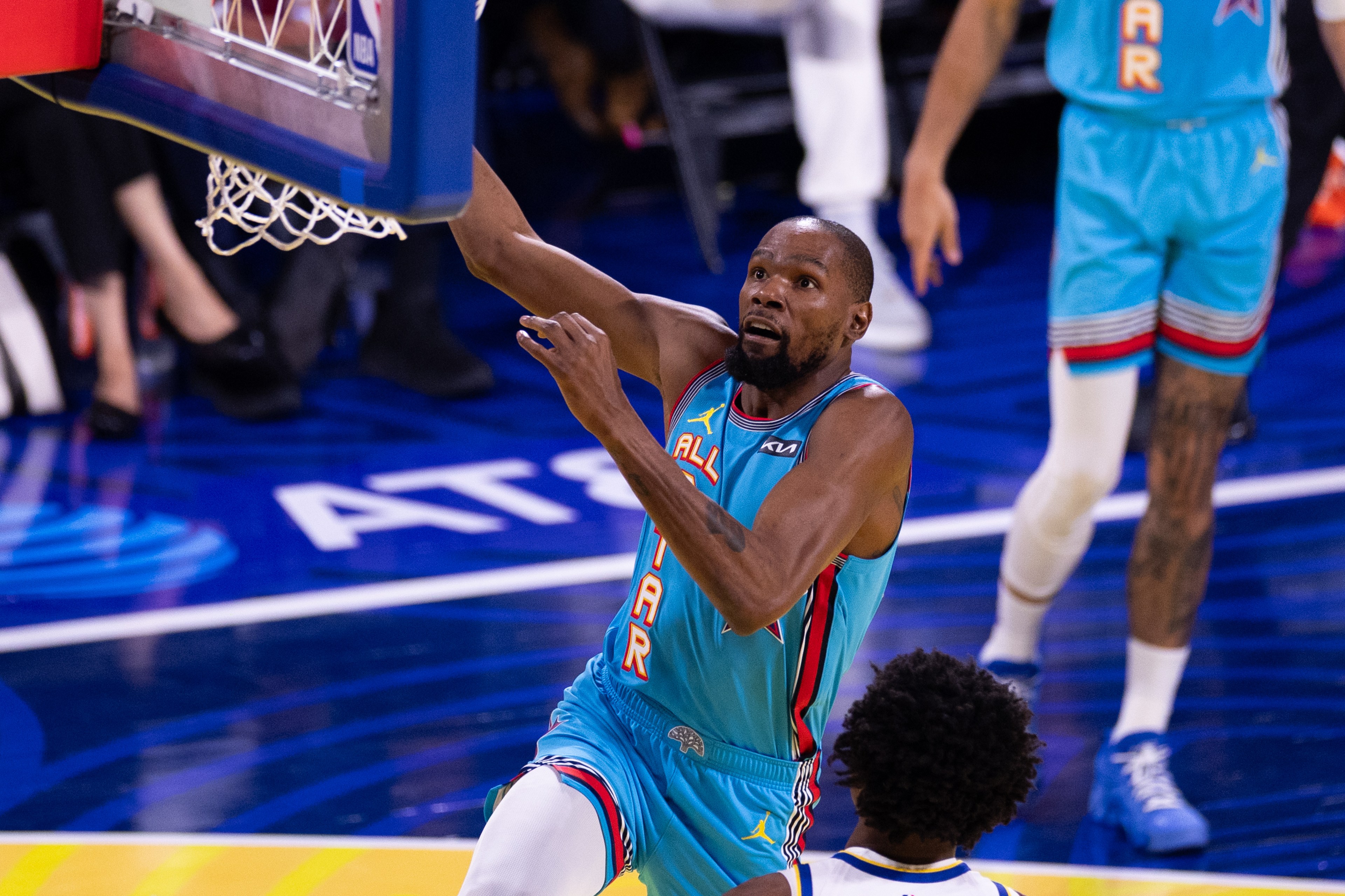 A basketball player in a blue &quot;All-Star&quot; jersey is making a layup, reaching towards the hoop, with other players visible in the background.