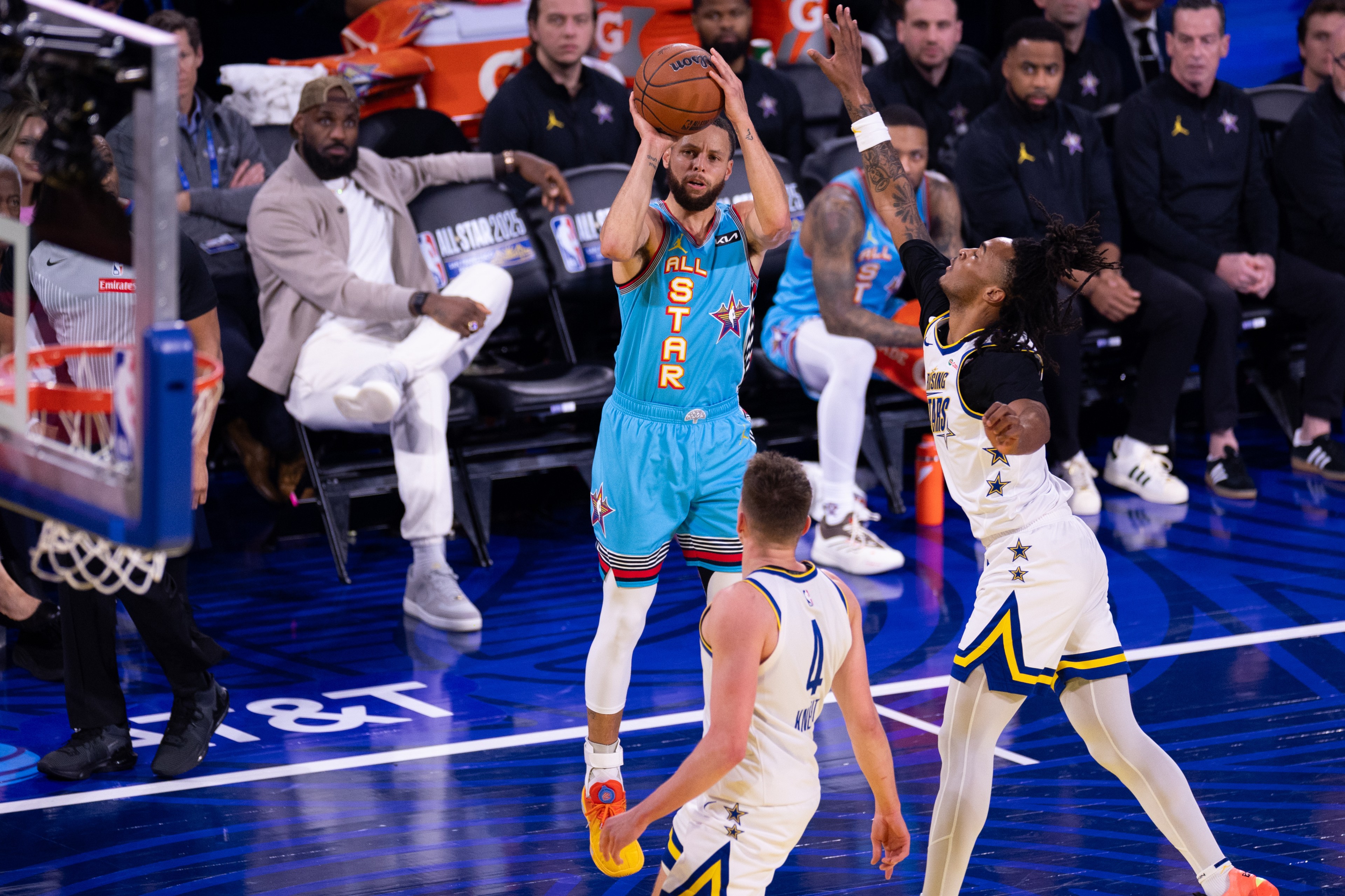 A basketball player in a teal &quot;All-Star&quot; uniform is shooting a jump shot while a defender in a white and black uniform tries to block the shot.