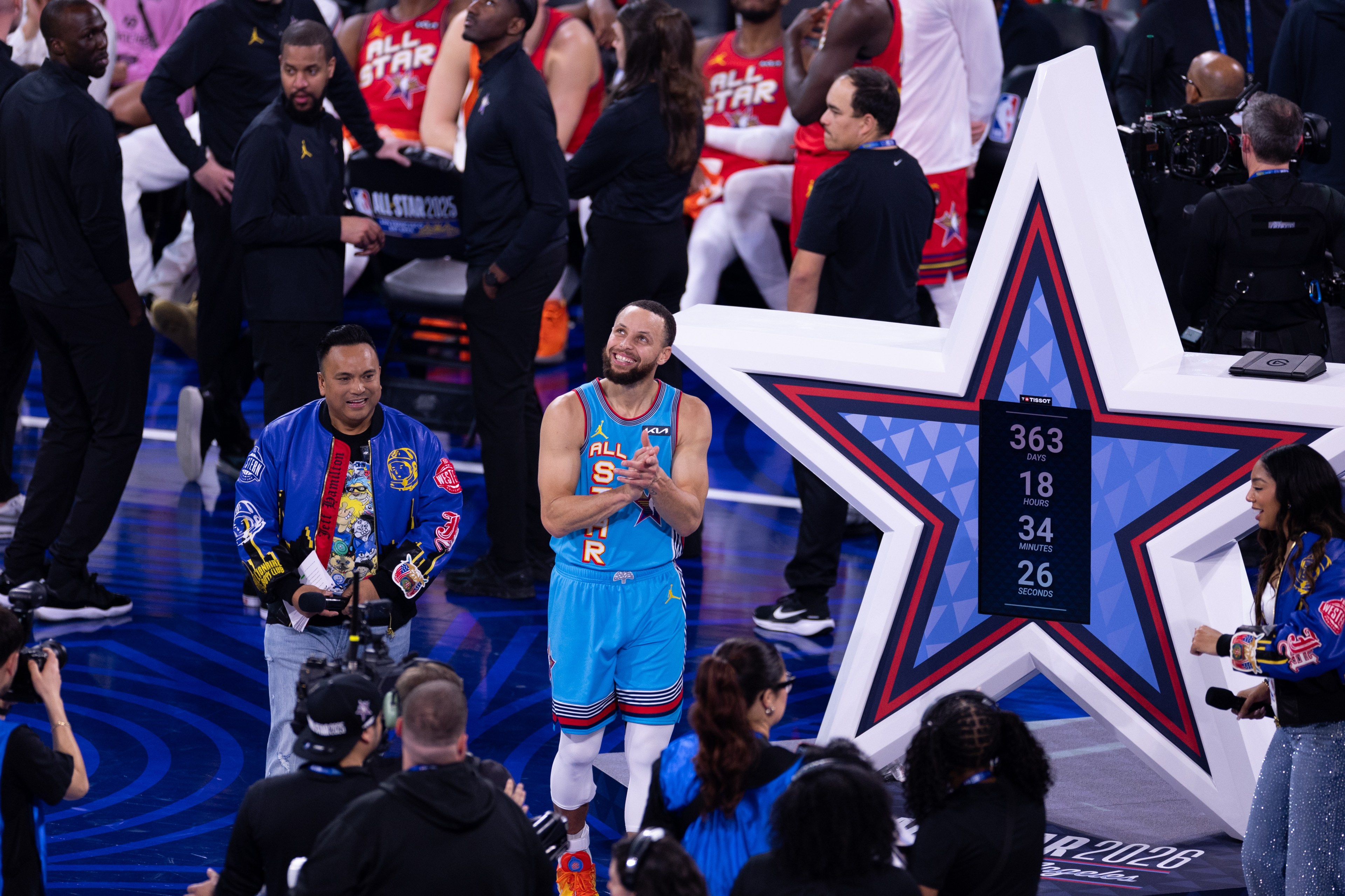 A smiling player in a blue &quot;All-Star&quot; jersey stands near a large star-shaped display counting down days, surrounded by people and camera crew on a basketball court.