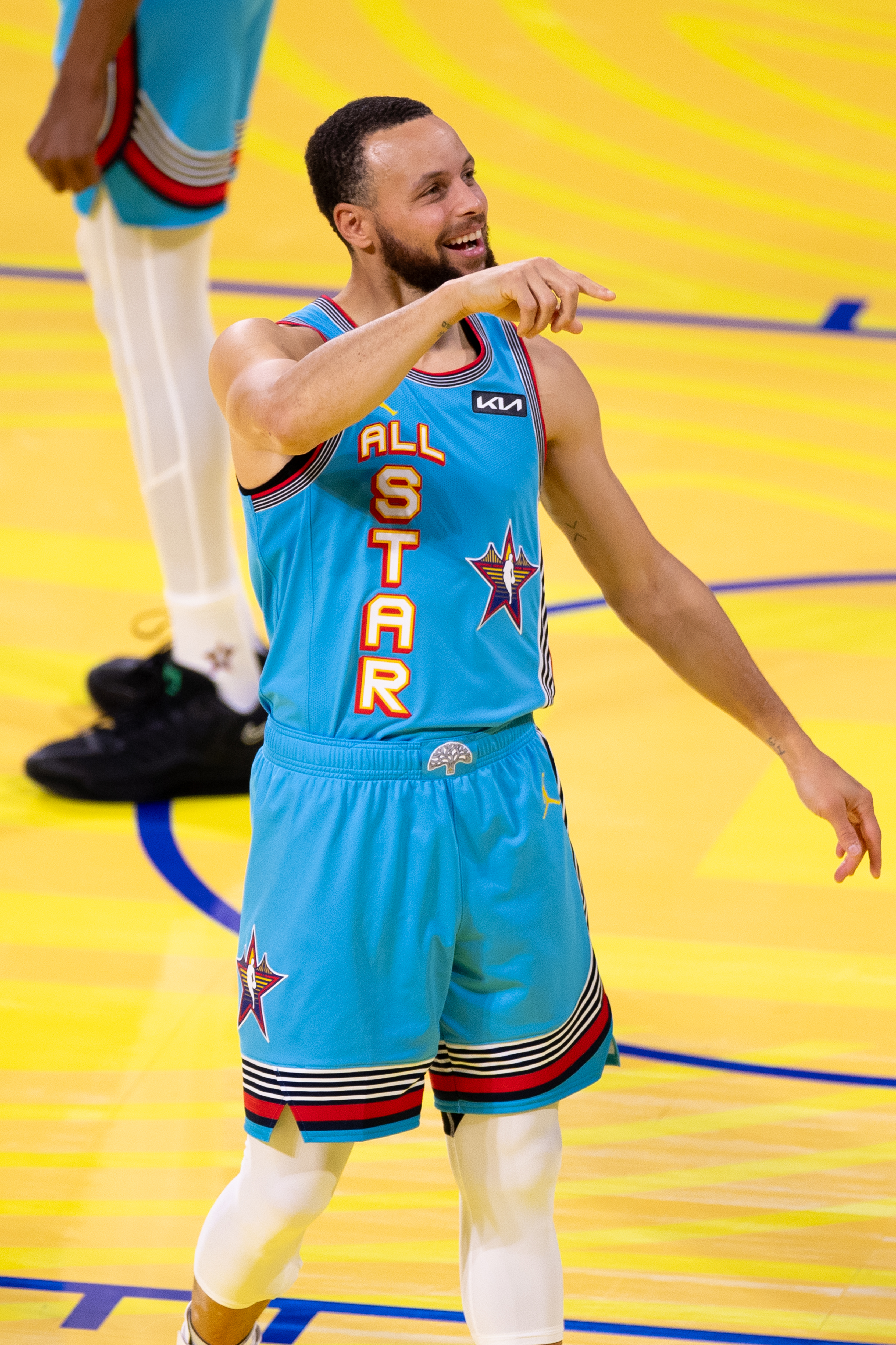 A basketball player wearing a light blue &quot;All Star&quot; uniform smiles and points while standing on a court with a yellow floor.