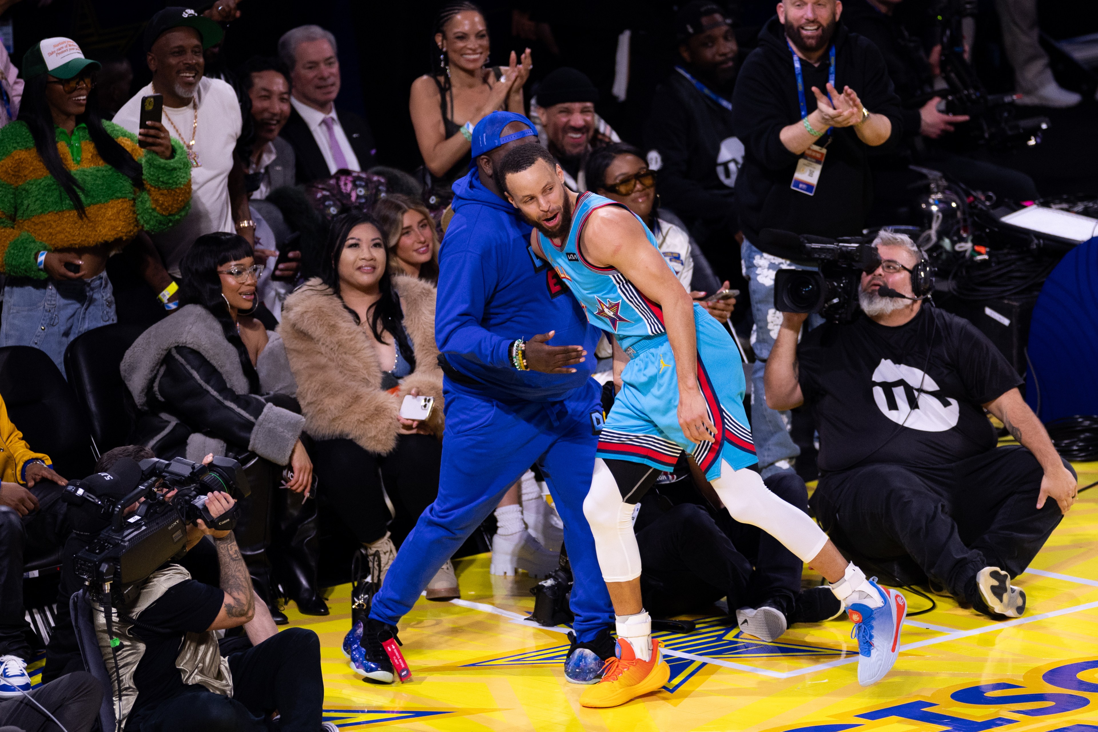 A basketball player in a blue uniform is interacting with a person in blue on the sidelines. People are cheering, and a cameraman is capturing the moment.