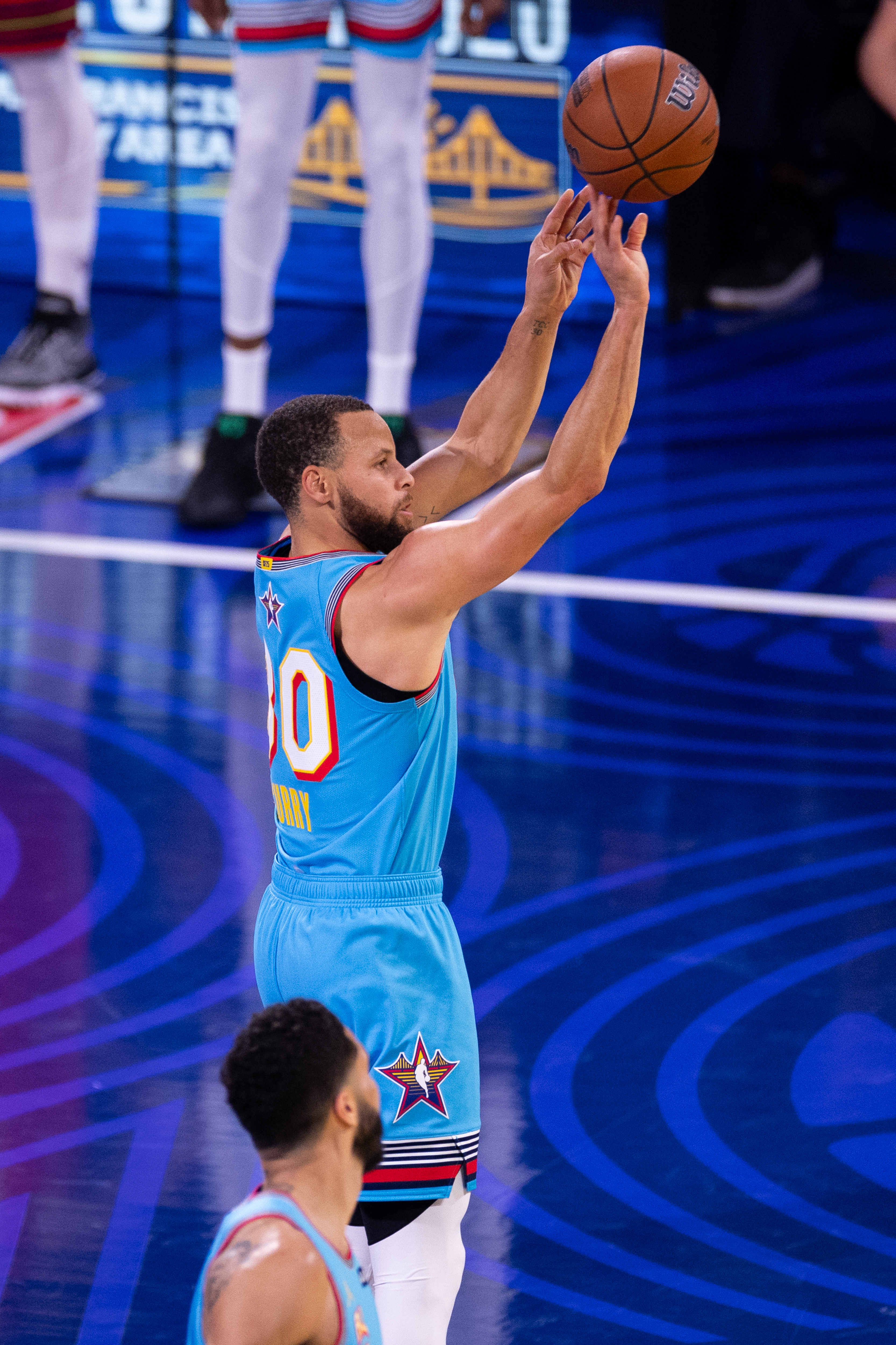 A basketball player in a blue jersey shoots a ball on a court. Another player is visible in the foreground, also wearing a similar blue jersey.