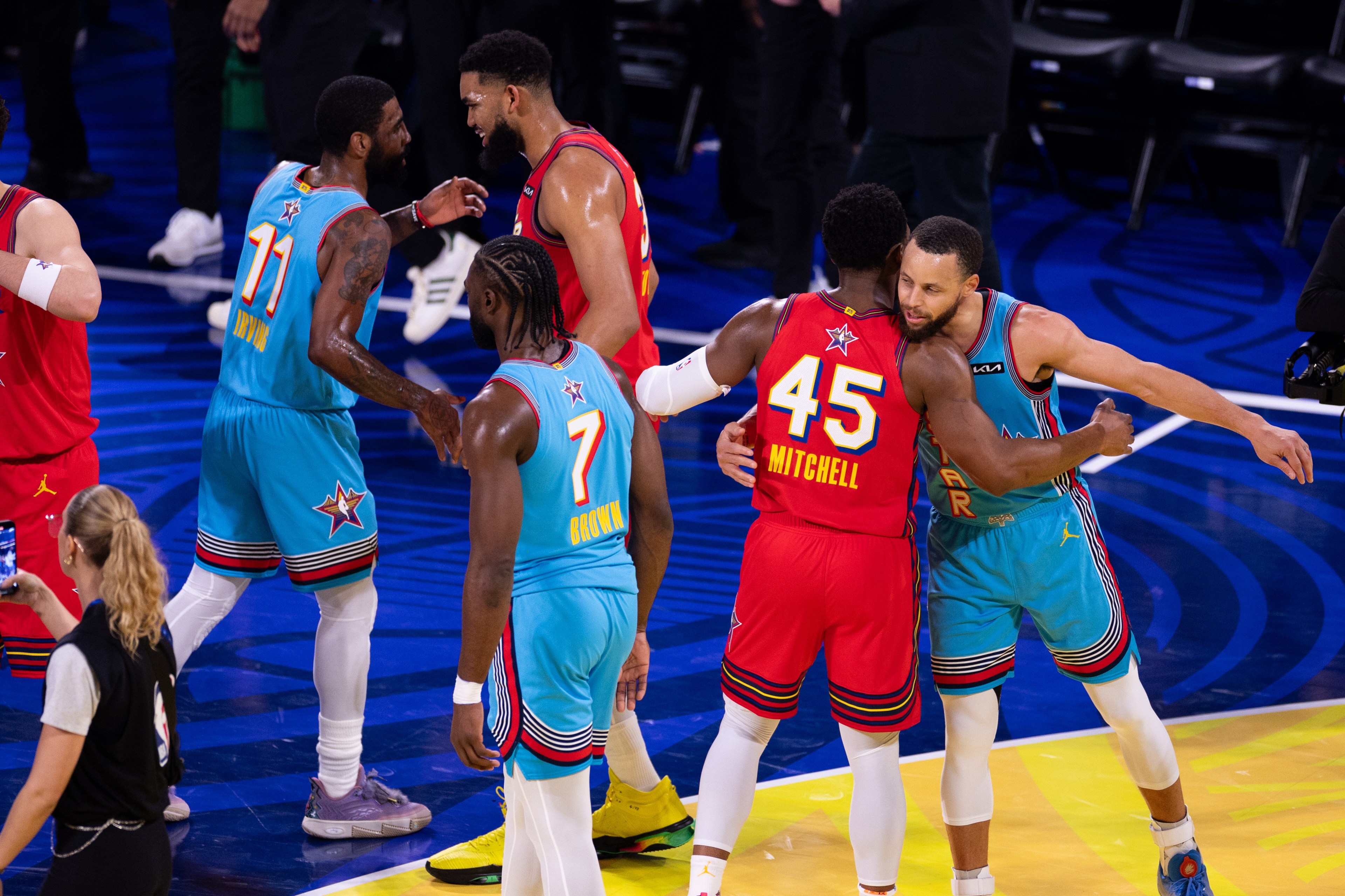 Basketball players in colorful uniforms embrace on the court, showing camaraderie after a game. Nearby, a woman stands with her back to the camera.