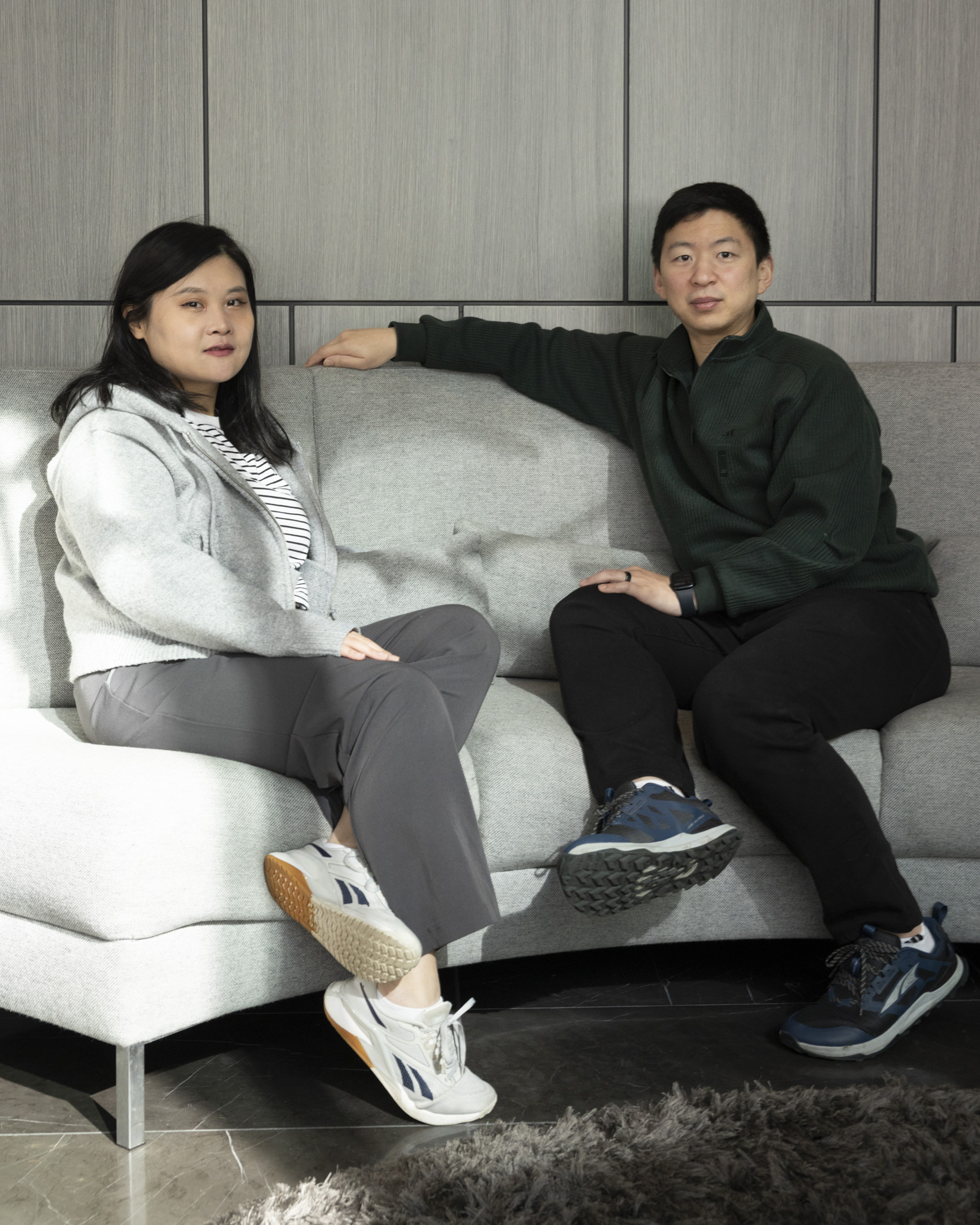 Two people sit on a gray sofa in a modern room with paneled walls. One wears a gray sweater and striped shirt, the other a dark sweater and pants. Both wear sneakers.