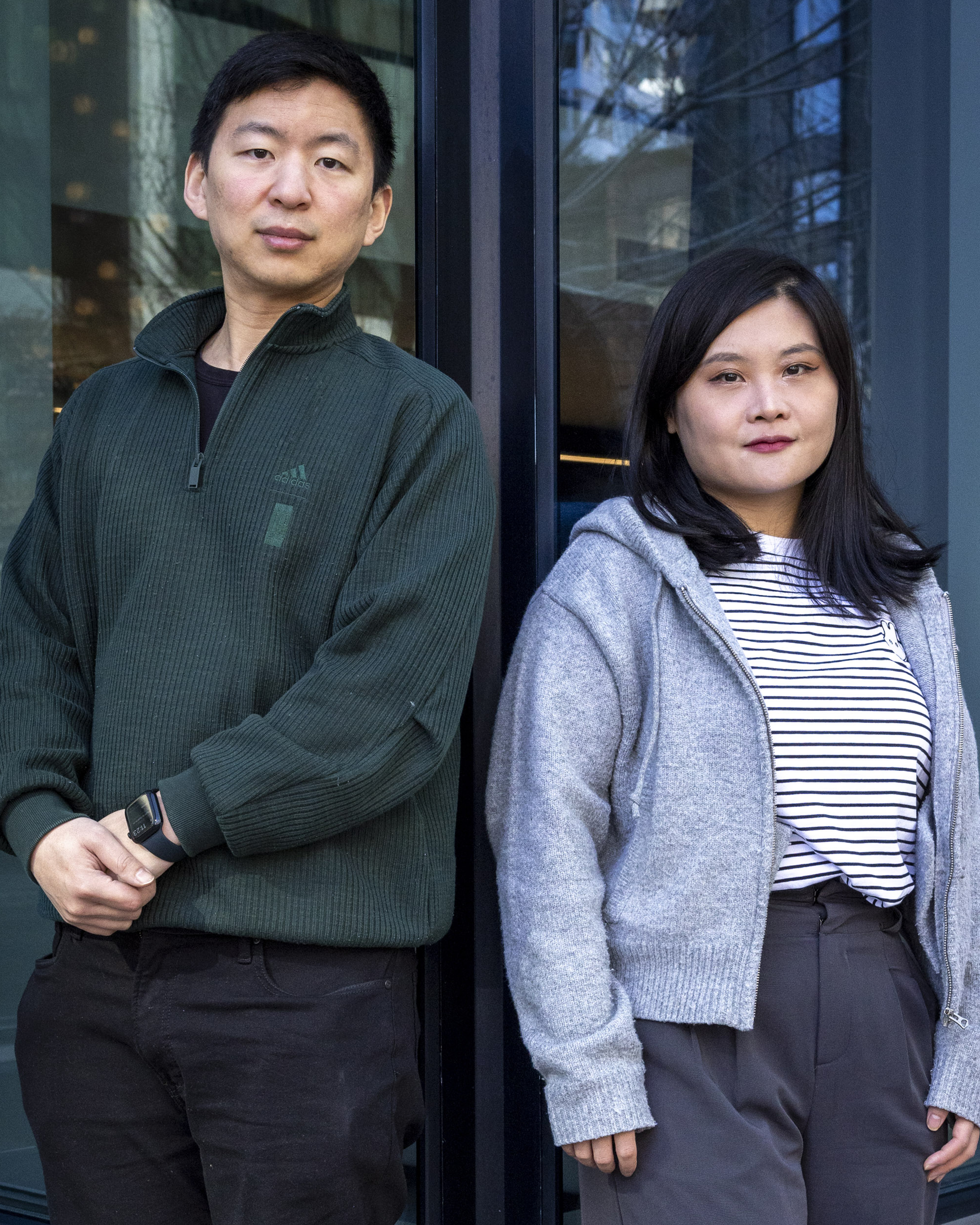 Two people stand side by side. One wears a dark green sweater, and the other a gray cardigan over a striped shirt. They are in front of a glass building.