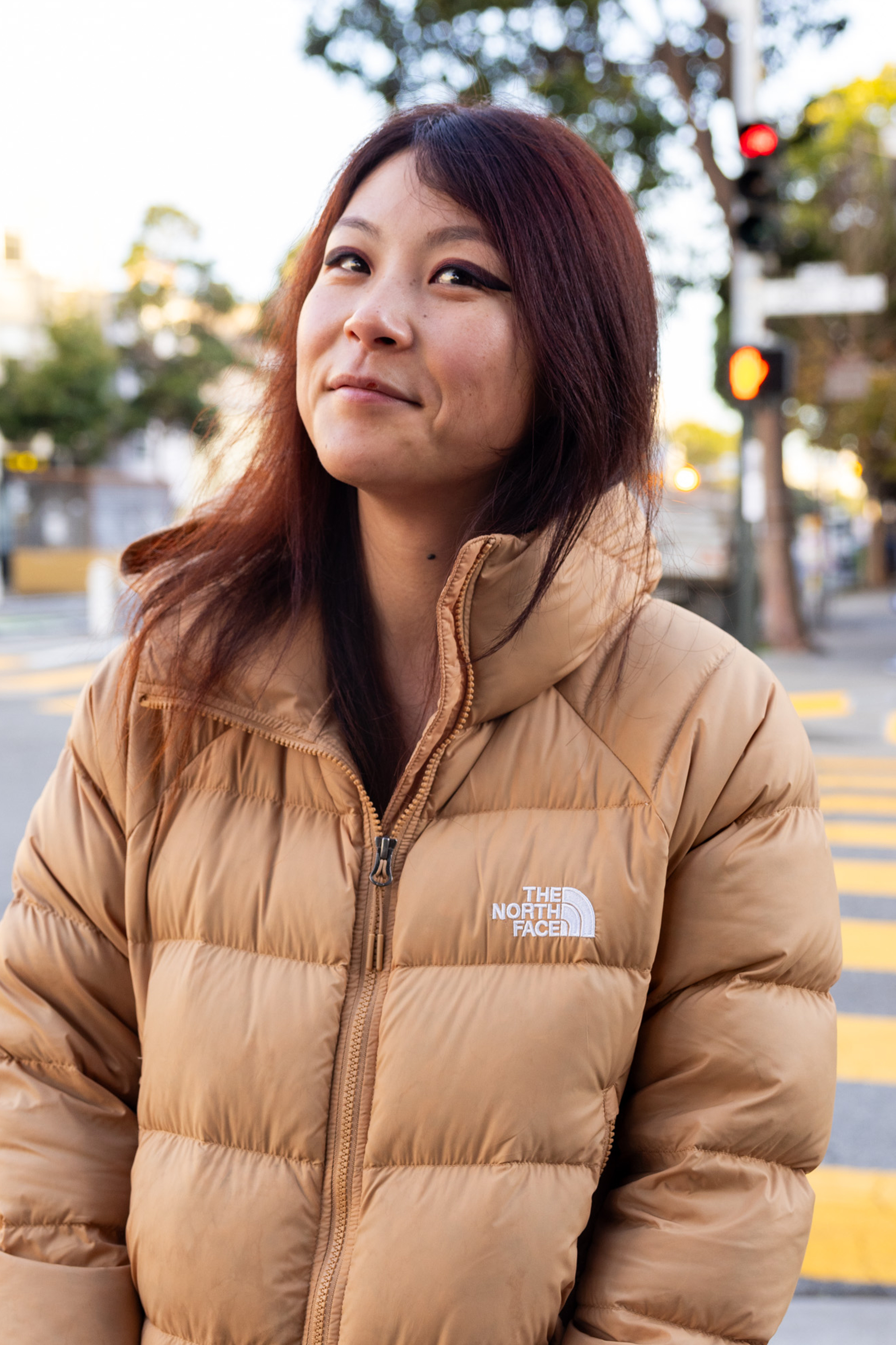 A woman with long hair smiles slightly while wearing a light brown puffer jacket. She stands outdoors near a crosswalk, and a traffic light glows red behind her.