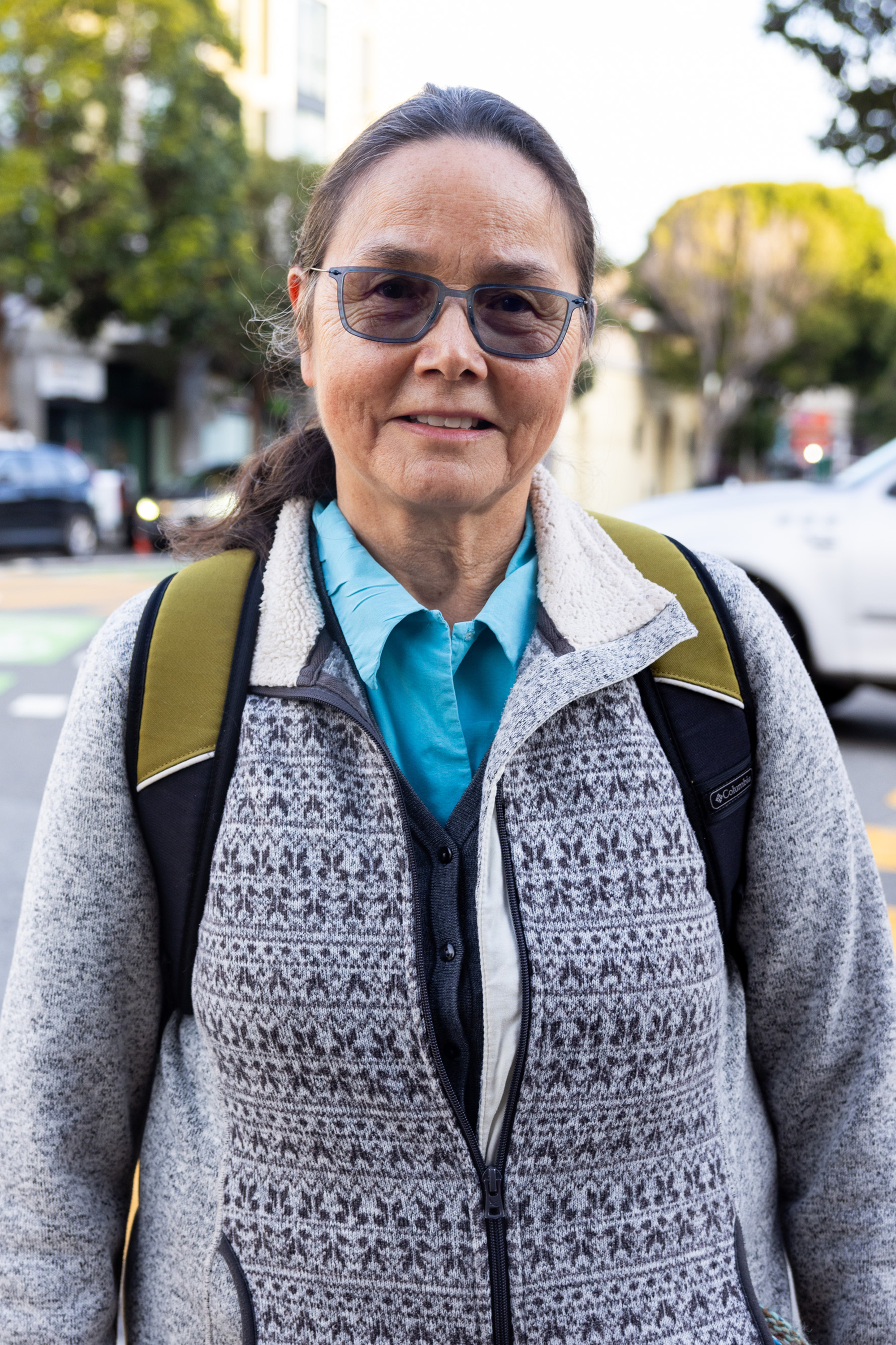 A person wearing glasses, a patterned sweater, and a backpack stands on a city street with trees and cars in the background.