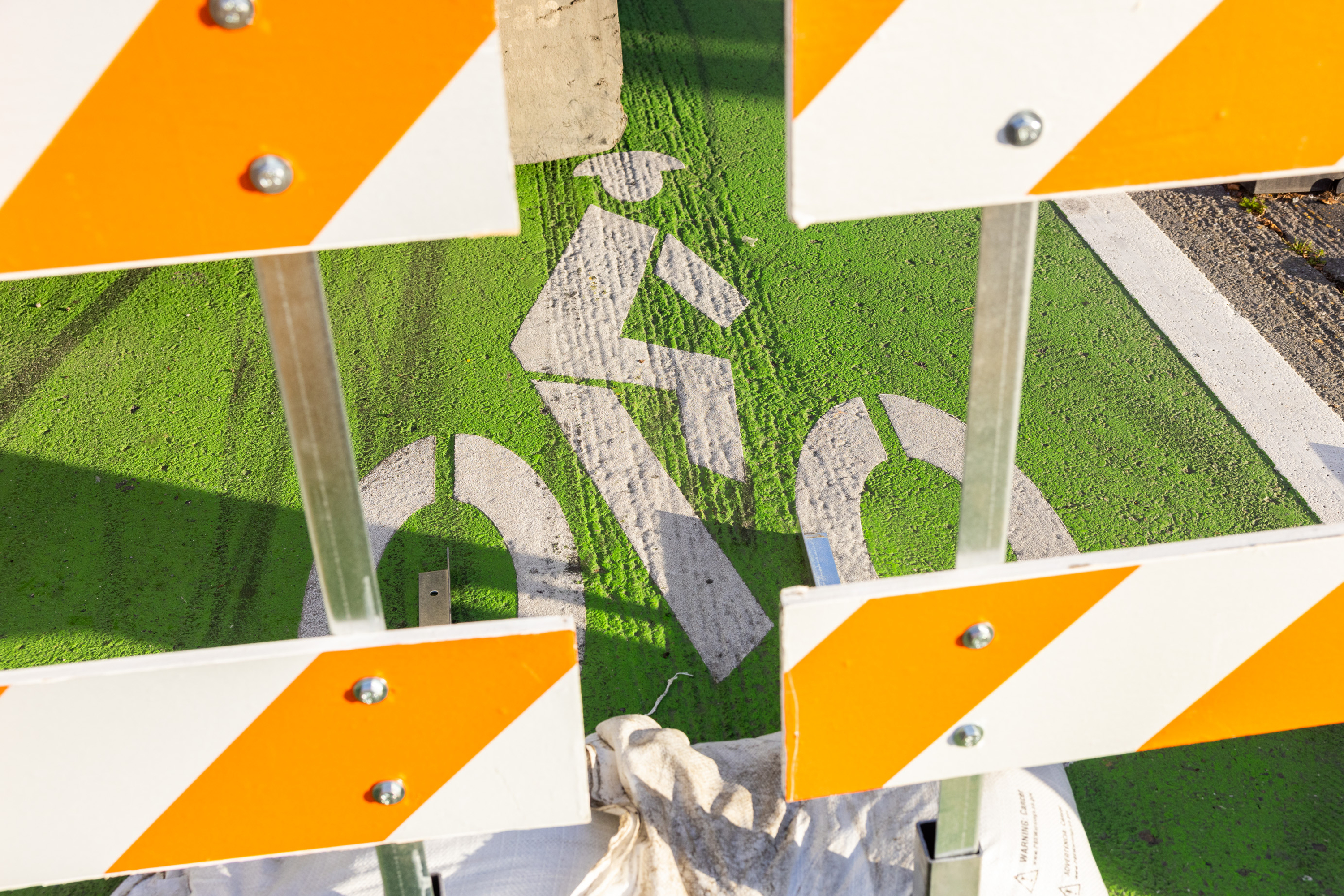 A road barrier with orange and white stripes partially obscures a green bike lane symbol on the ground, indicating a path for cyclists.