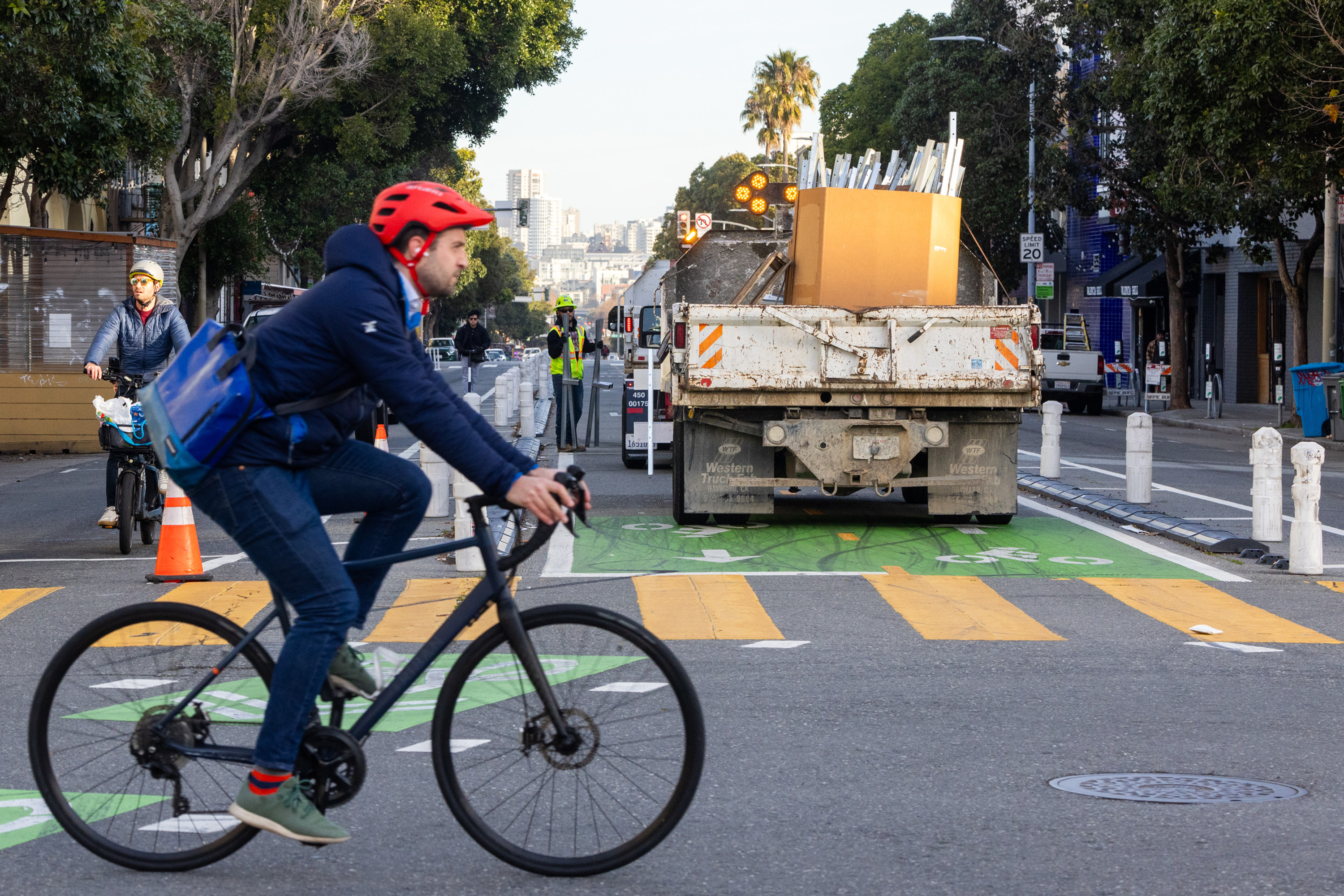 A cyclist in a red helmet rides past a truck loaded with items on a city street. Another cyclist approaches from the opposite direction. Trees line the street.