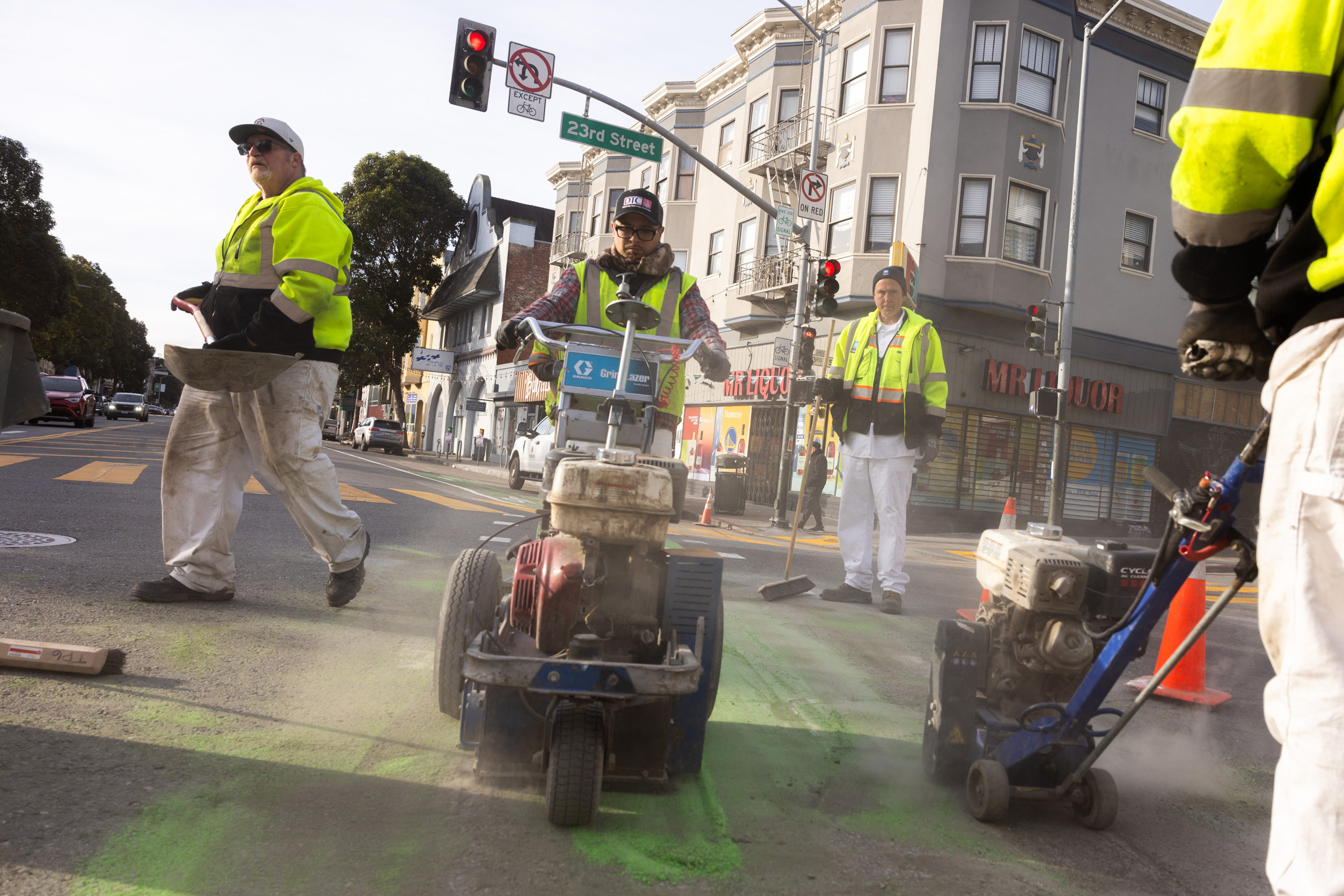 Workers in yellow vests use machines to paint green markings on a city street. They are near an intersection with buildings and traffic lights.