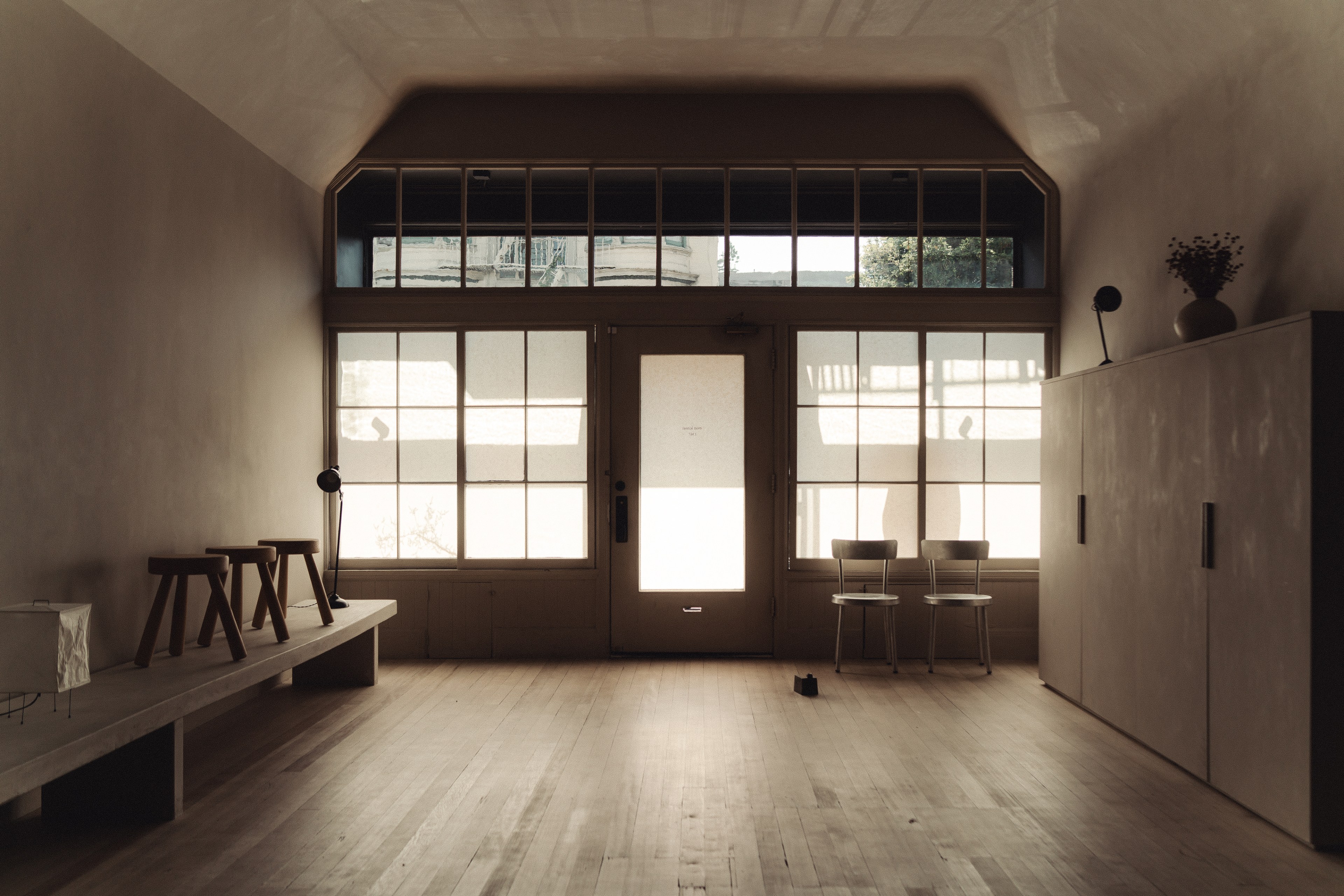 The image shows a minimalist room with wooden floors, stools, and chairs by a large windowed door, softly lit with natural light. A vase sits on a cabinet.