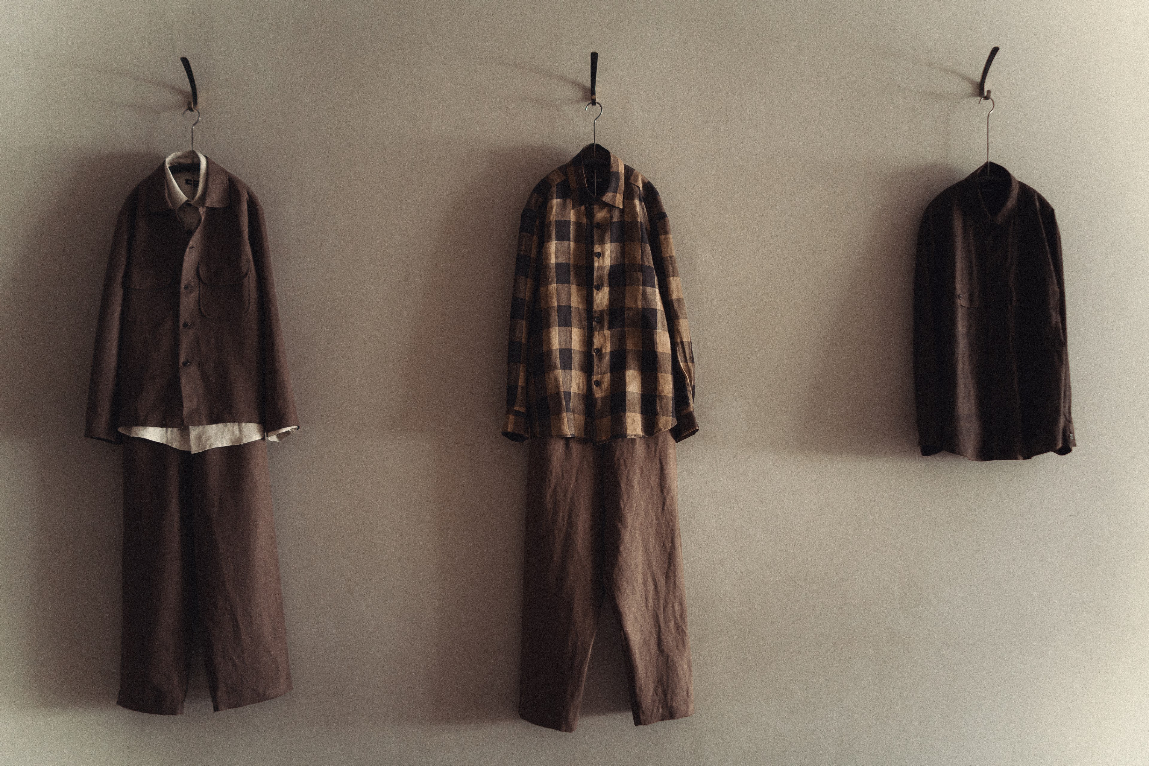 Three outfits hang on a wall: a brown jacket and pants, a checkered shirt with brown pants, and a dark button-up shirt.