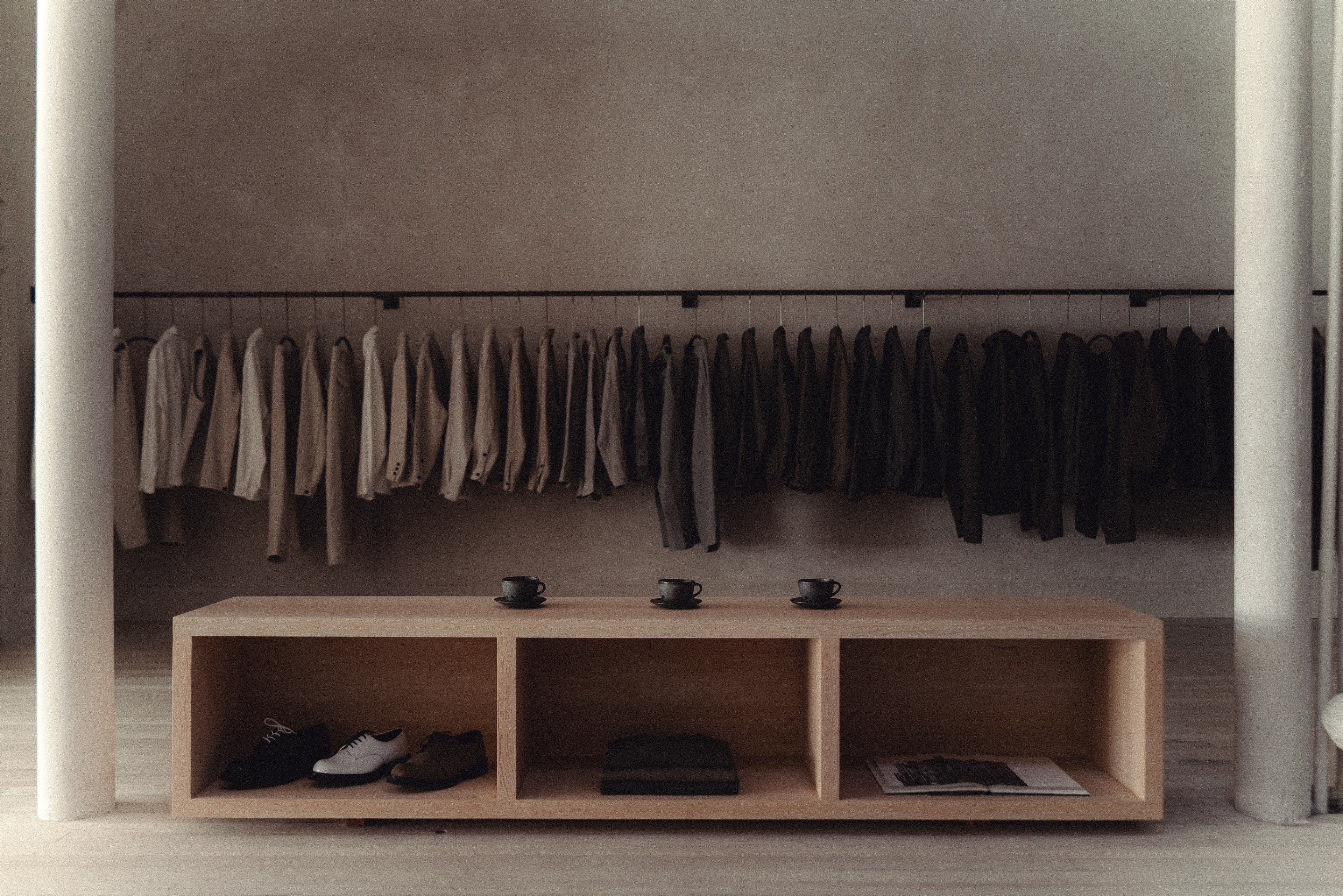 A minimalist clothing store features neatly hung jackets in neutral tones, a wooden display table with three cups on top, and shoes and books underneath.