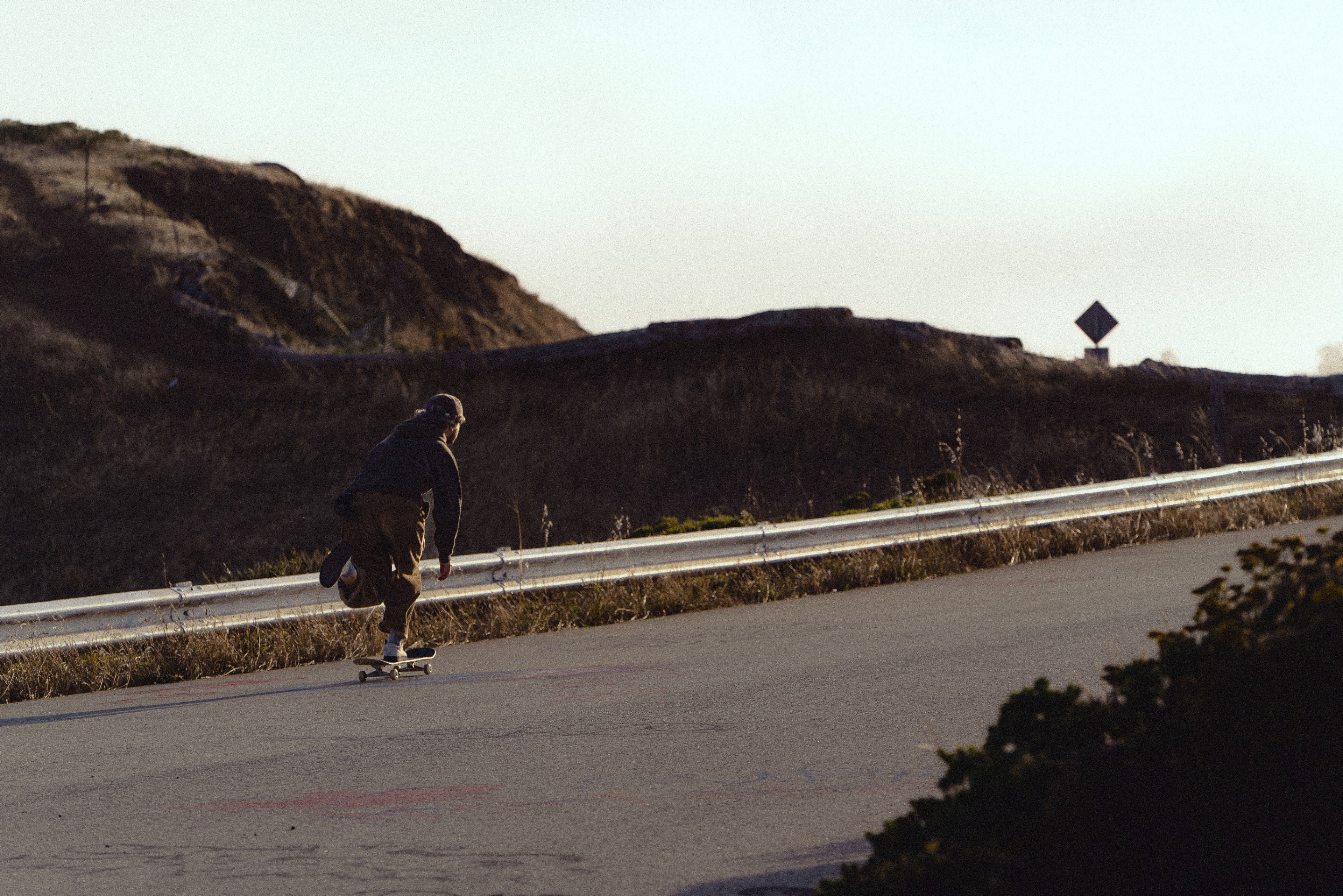 A person skateboards down a paved road beside a metal guardrail. The landscape features grassy hills and a road sign in the distance under a bright sky.