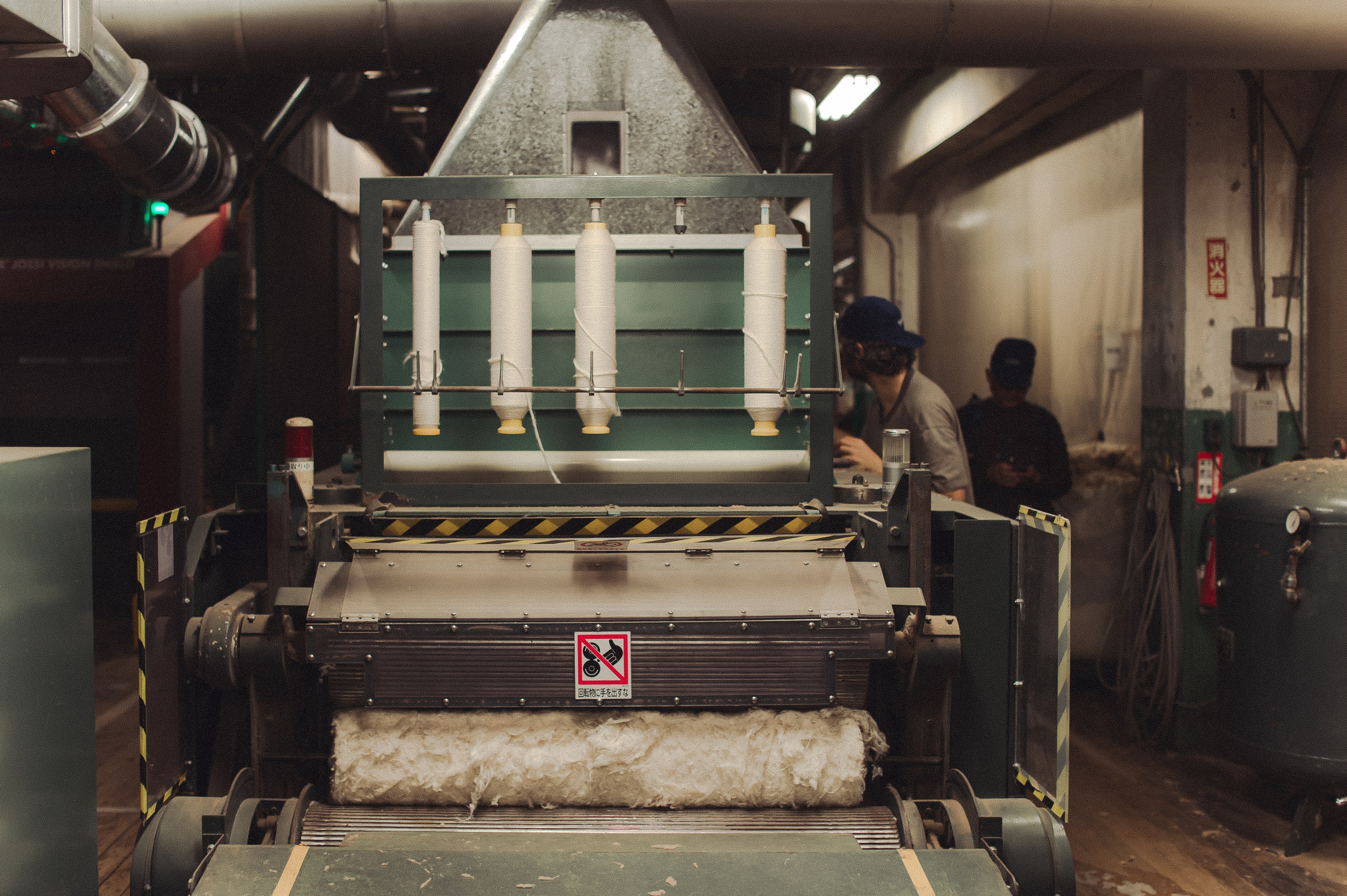 The image shows a machine in a factory with cotton or wool being processed. Spools of thread hang above, and two people are working in the background.