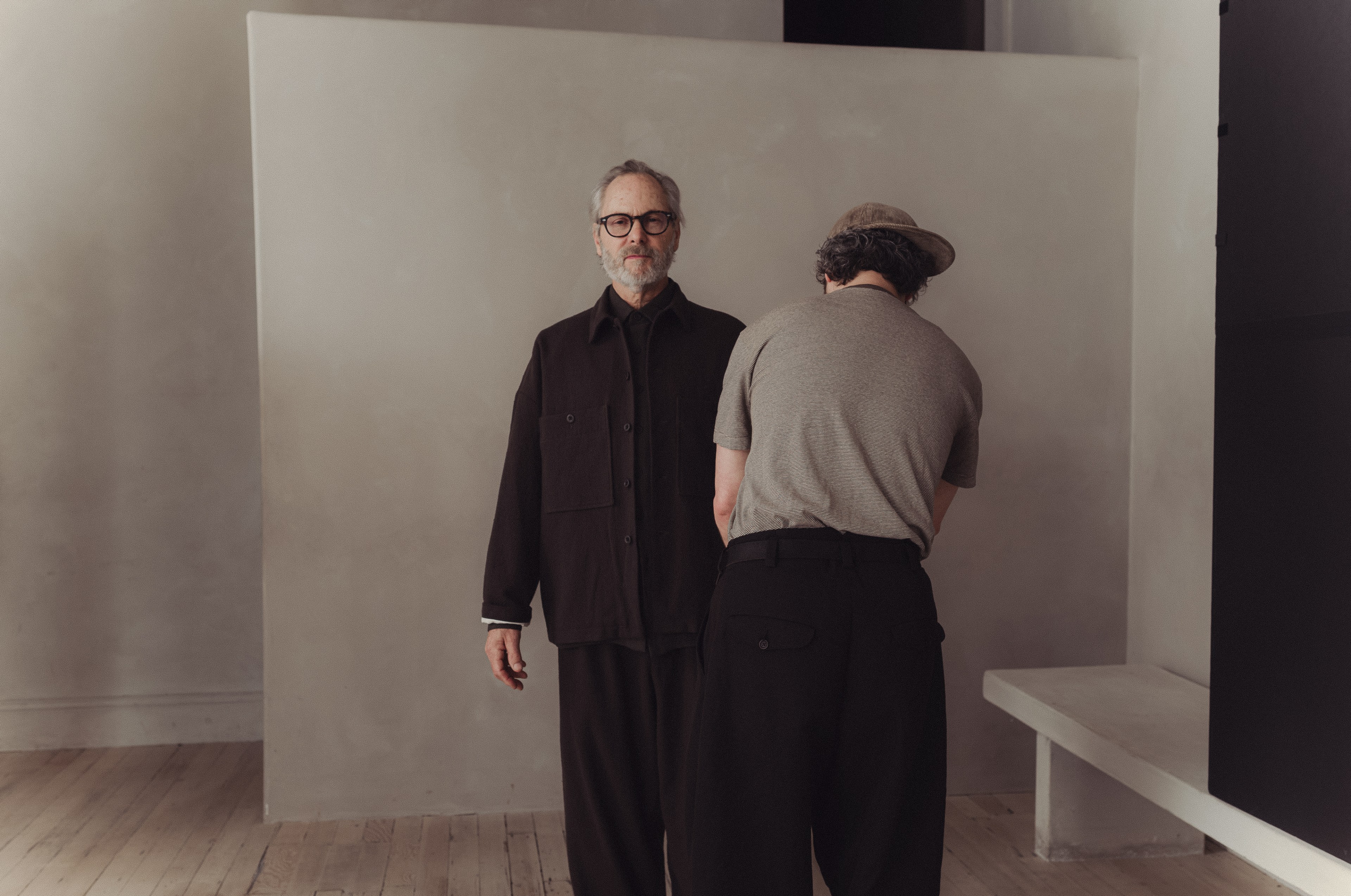 Two men stand in a minimalist room; one faces forward in dark clothing, while the other faces away, wearing a hat and gray shirt.