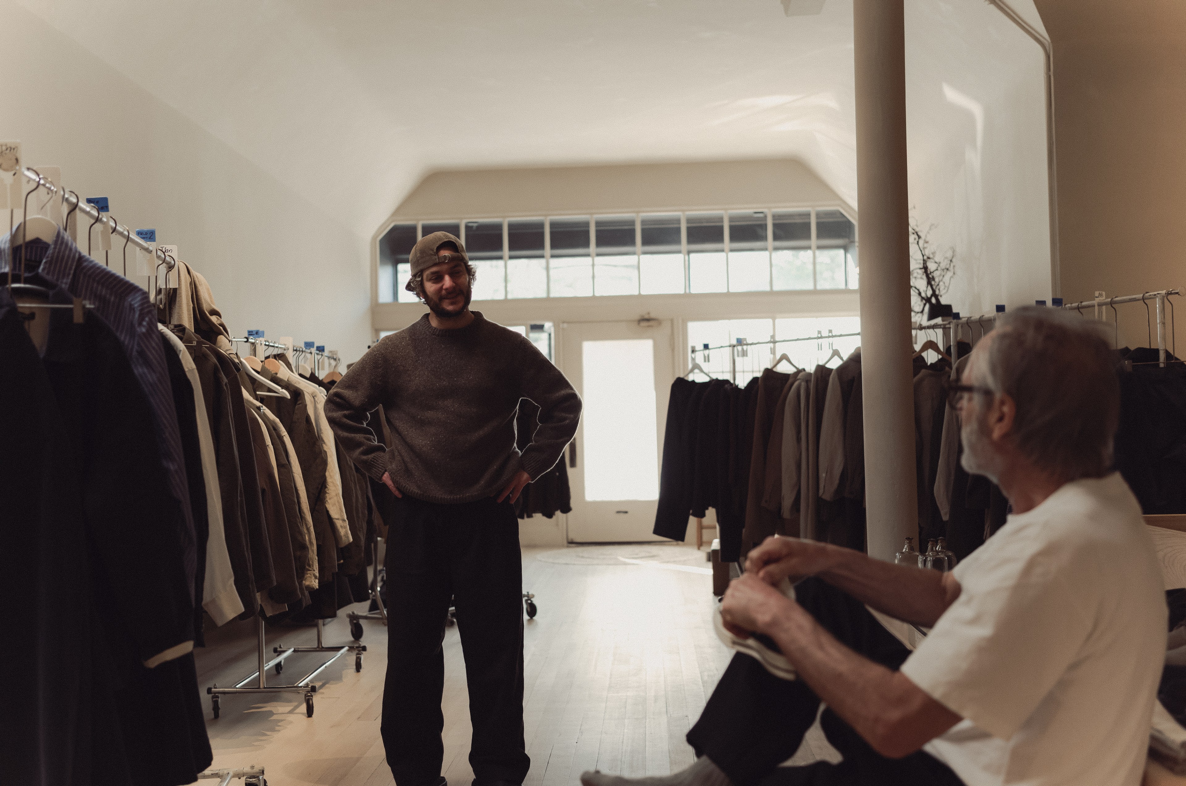 A man in a cap stands with hands on hips in a clothing store, while another person sits in the foreground. Racks of clothes line the walls under soft lighting.