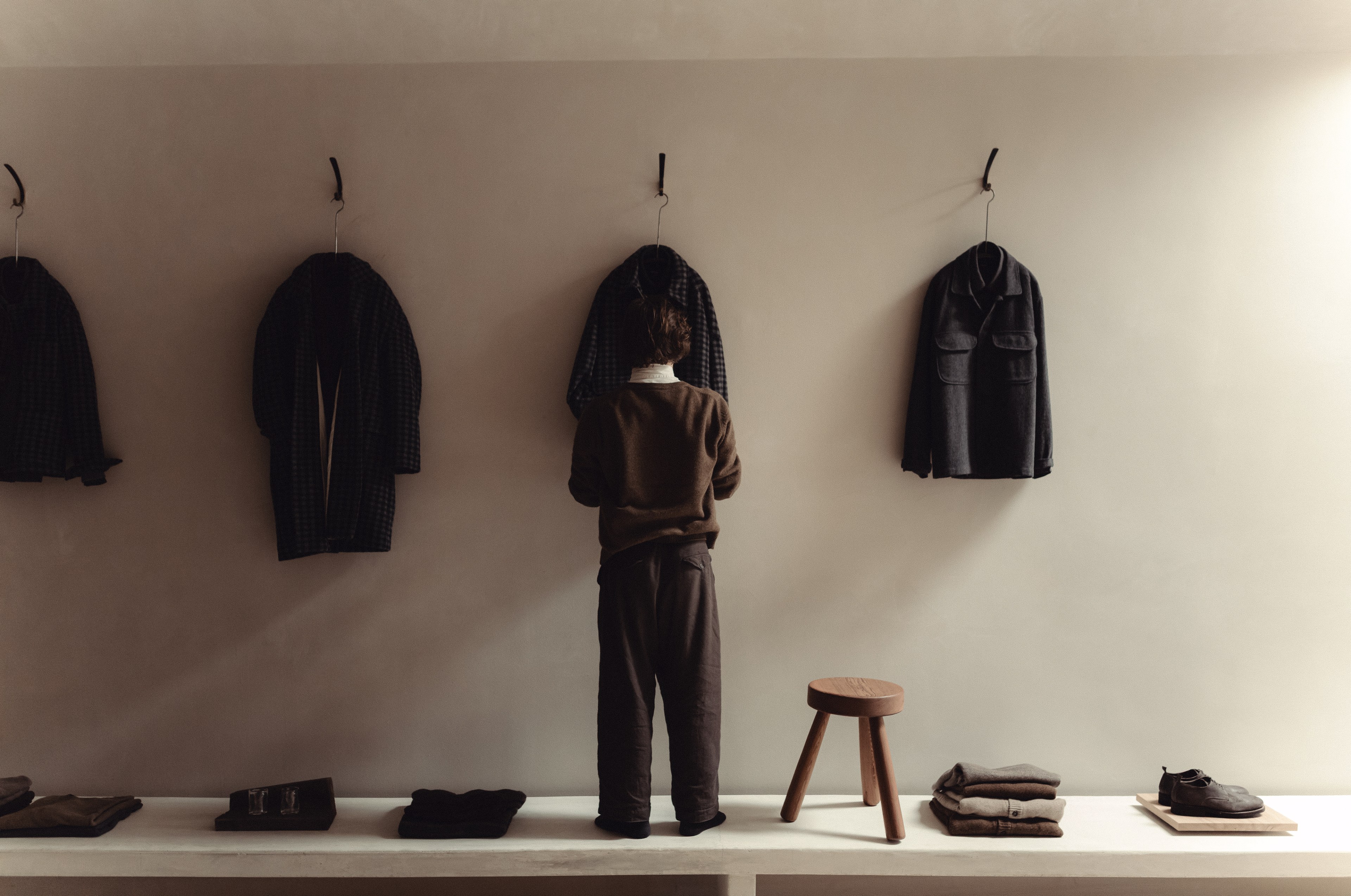 A person stands in a minimalistic room facing a wall displaying three dark coats. Below, clothes, shoes, and a small wooden stool are arranged neatly.