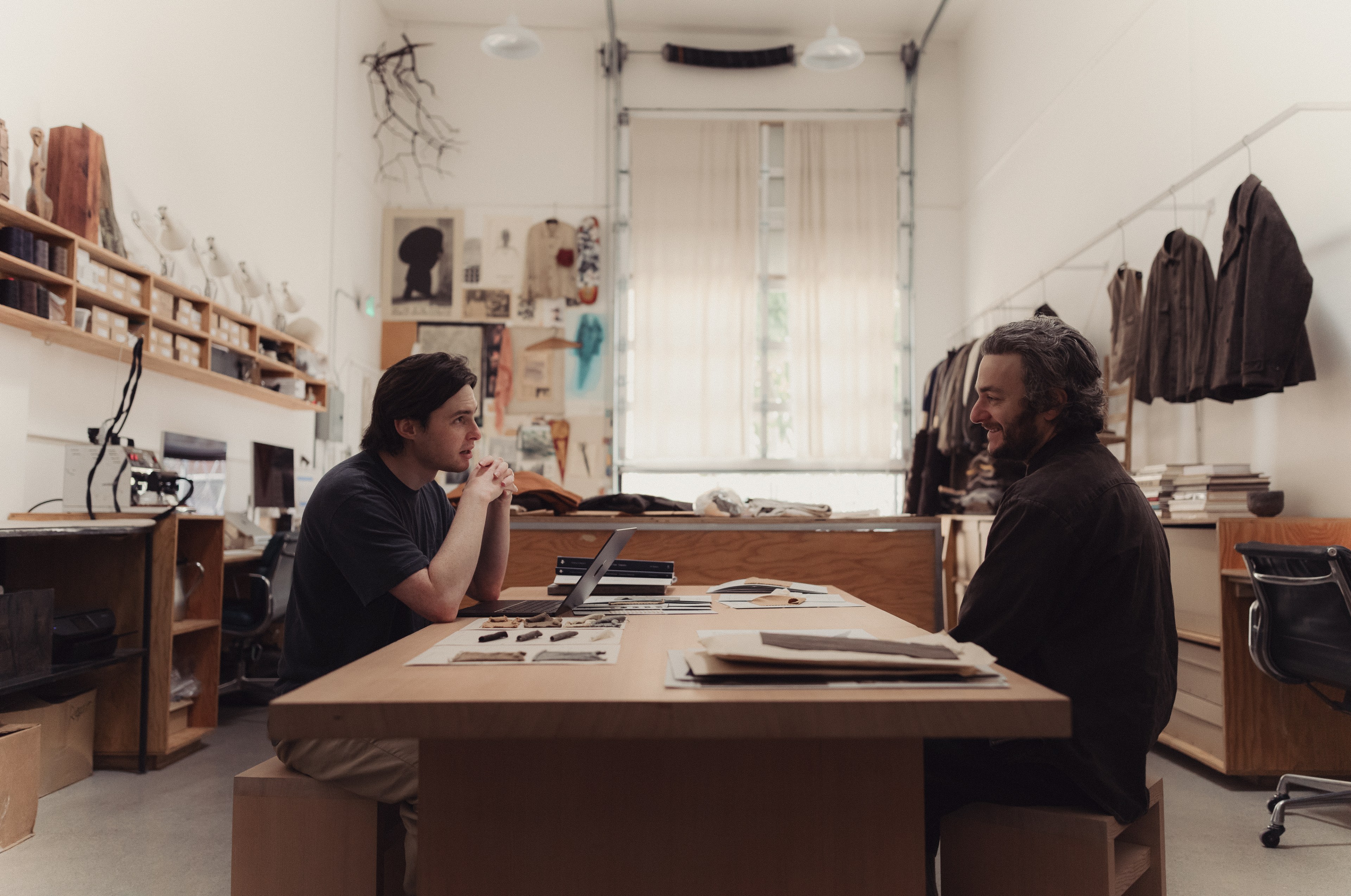 Two people are seated at a wooden table in a creative studio, surrounded by papers and fabrics. The space is bright, with artwork and clothing on the walls.