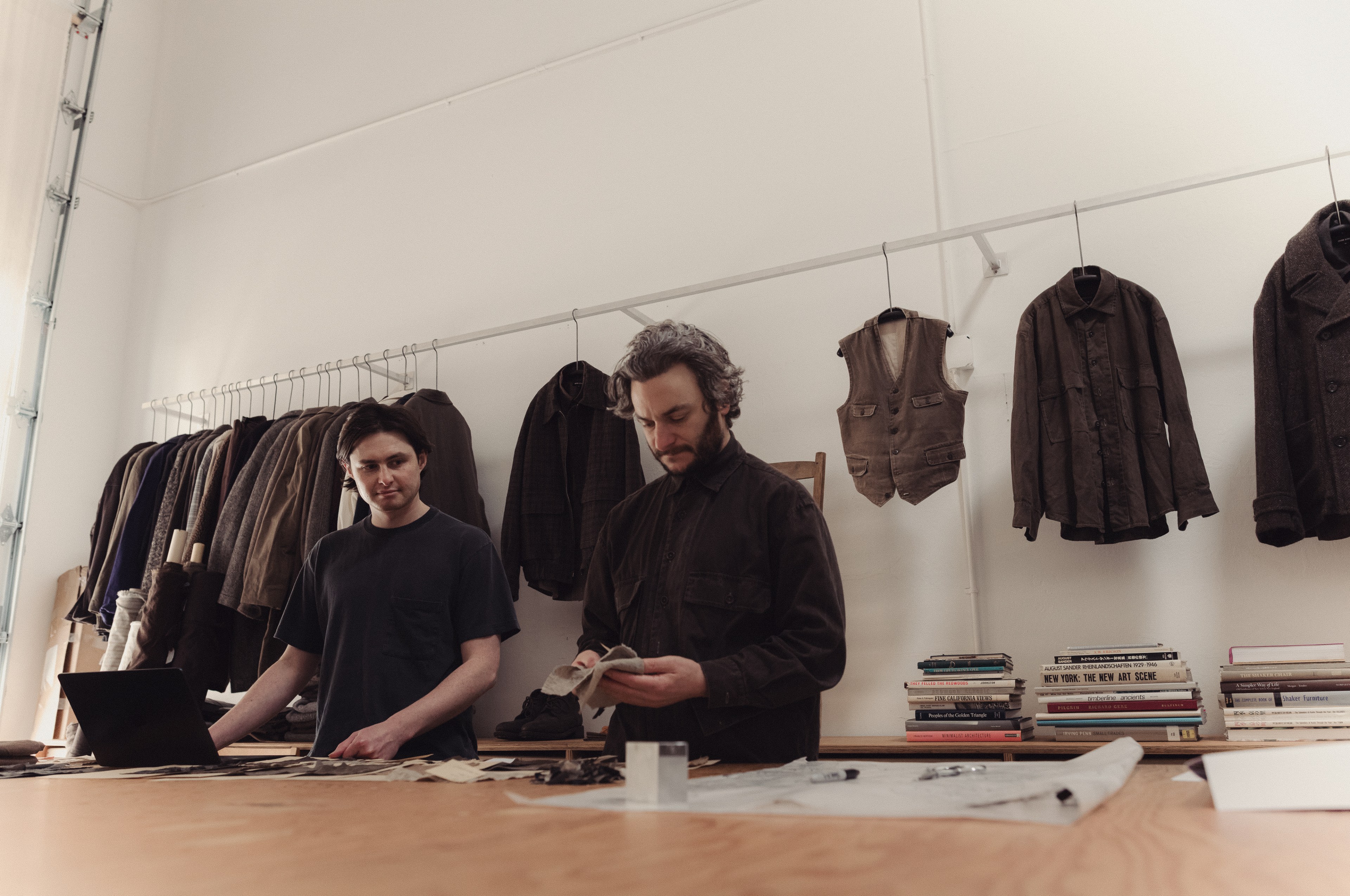 Two men are in a clothing workshop with fabric samples on a table. Behind them, racks hold jackets and vests. Books are stacked on a shelf.