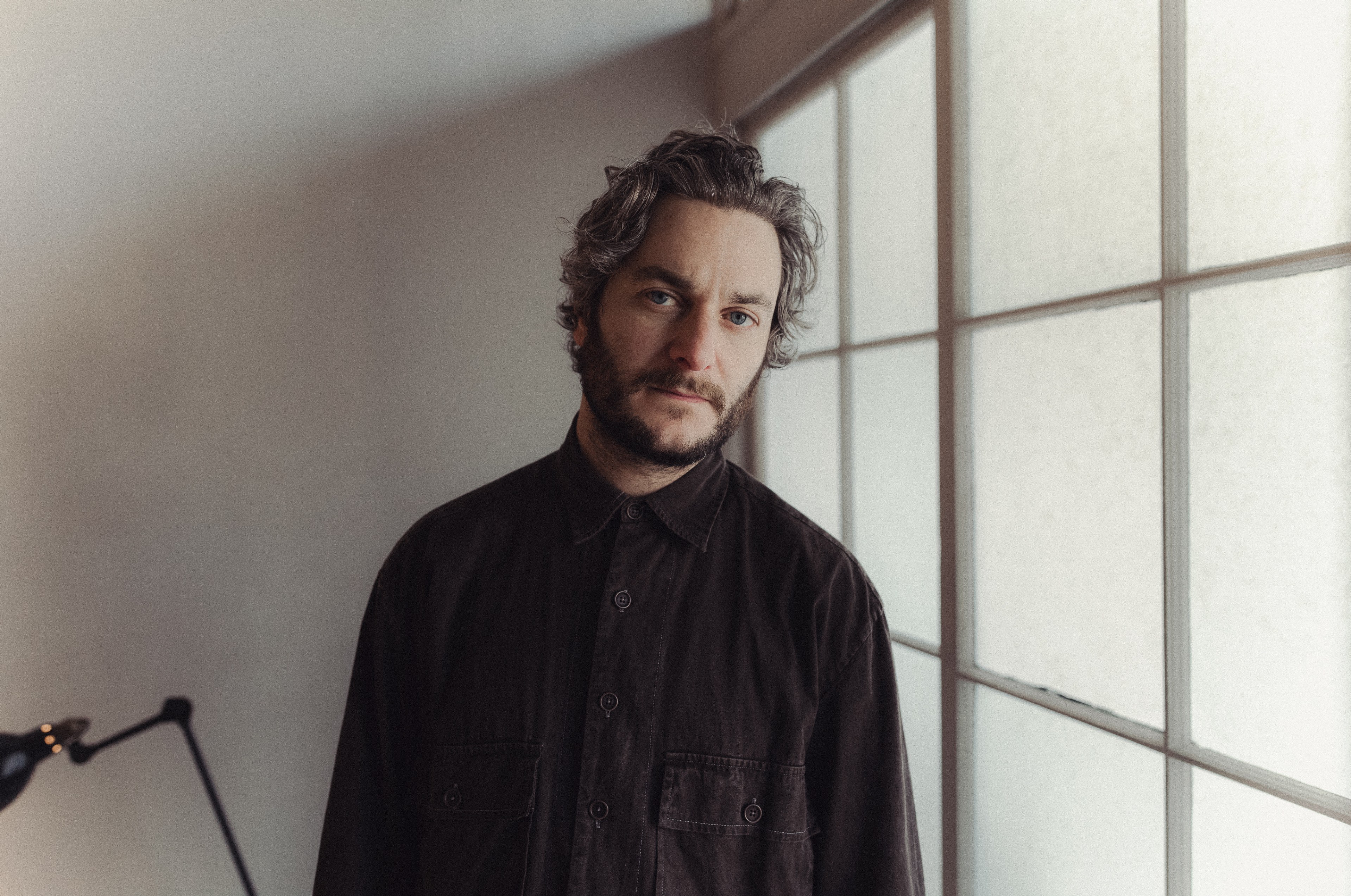 A man with wavy hair and a beard wears a dark shirt, standing near large windows in a softly lit room.