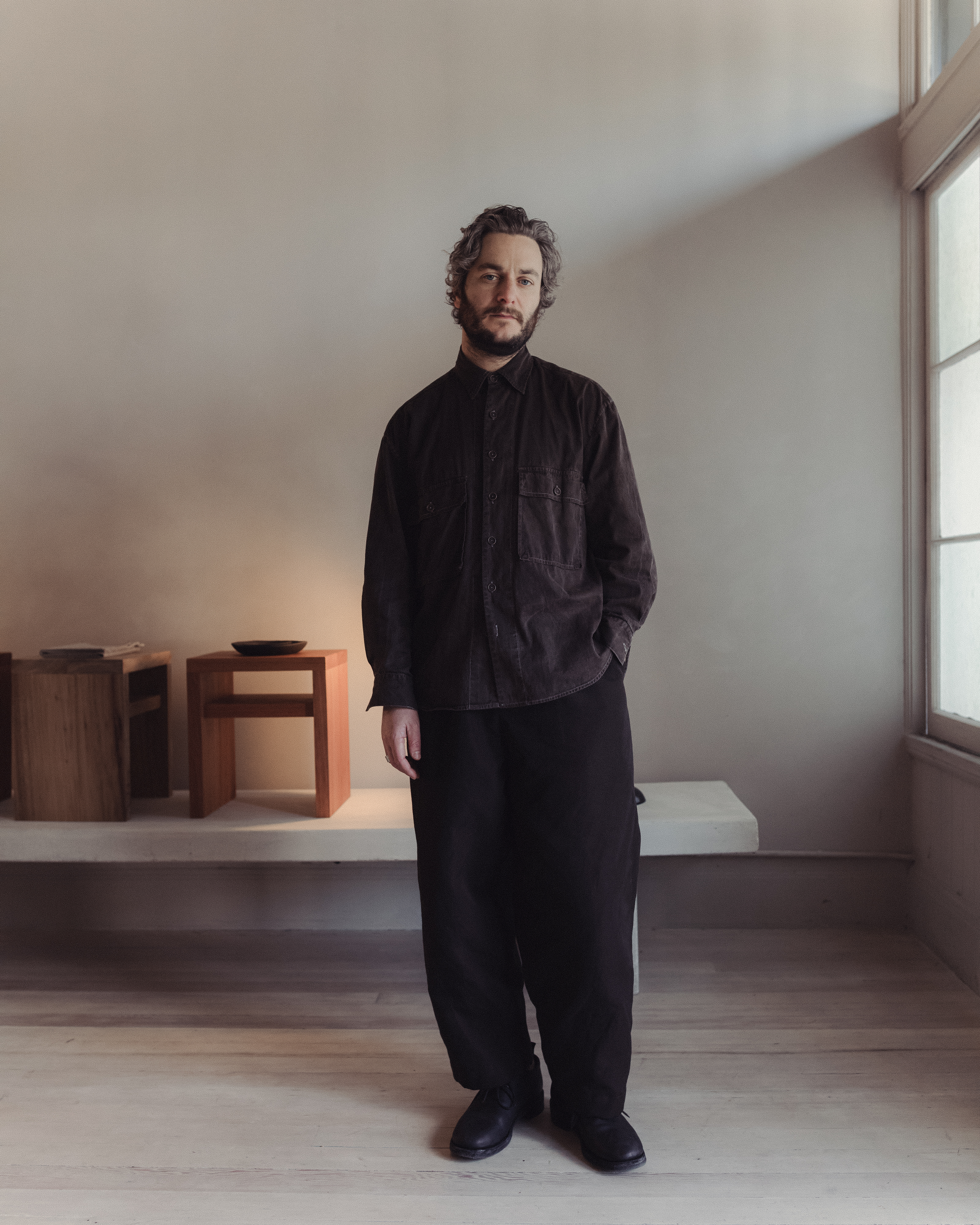 A man stands indoors wearing dark, loose-fitting clothes. The room is minimally decorated with wooden furniture and soft, natural lighting.