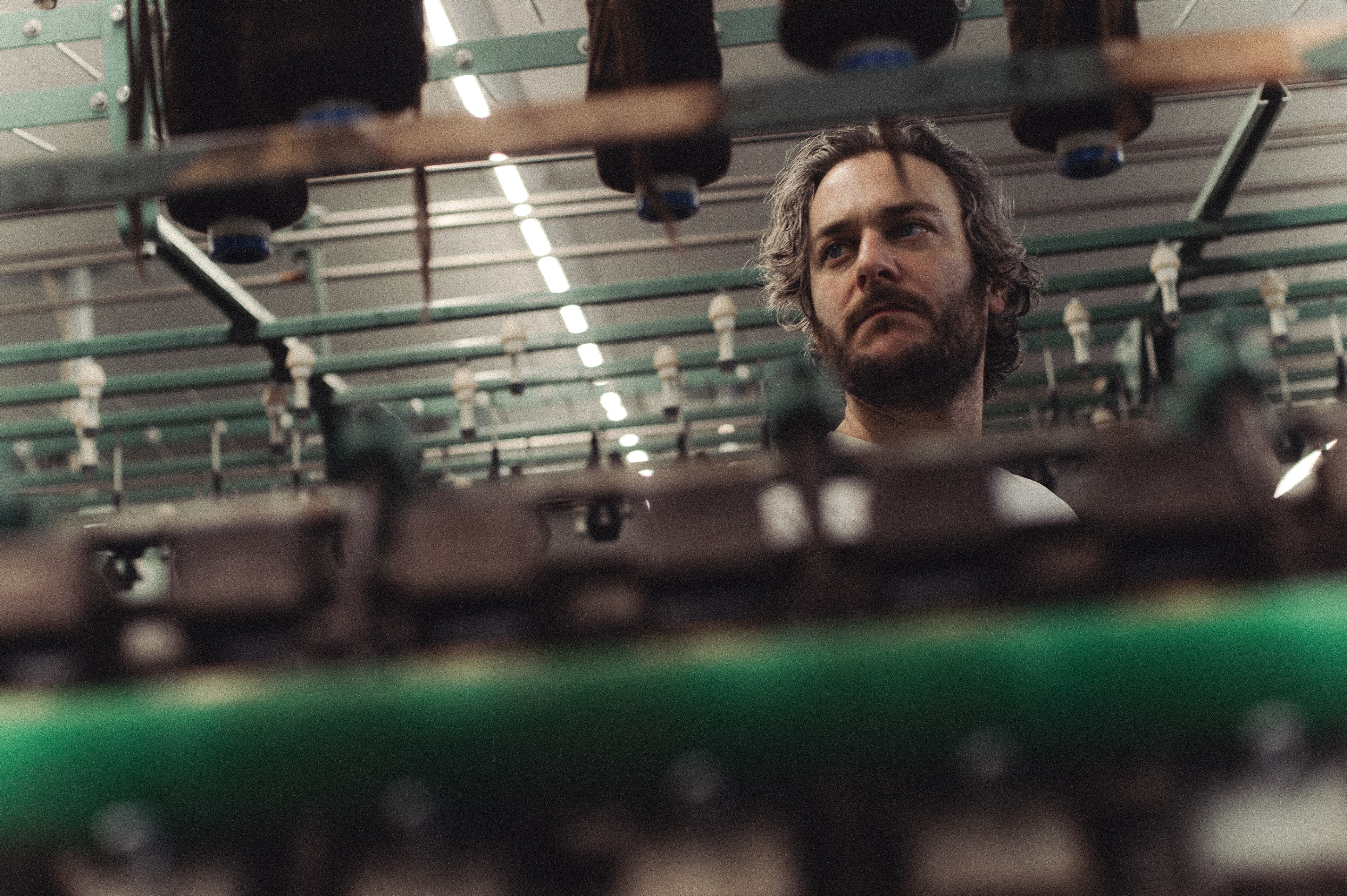 A bearded man is seen amidst a complex array of industrial machinery, appearing focused and contemplative. The setting suggests a manufacturing environment.