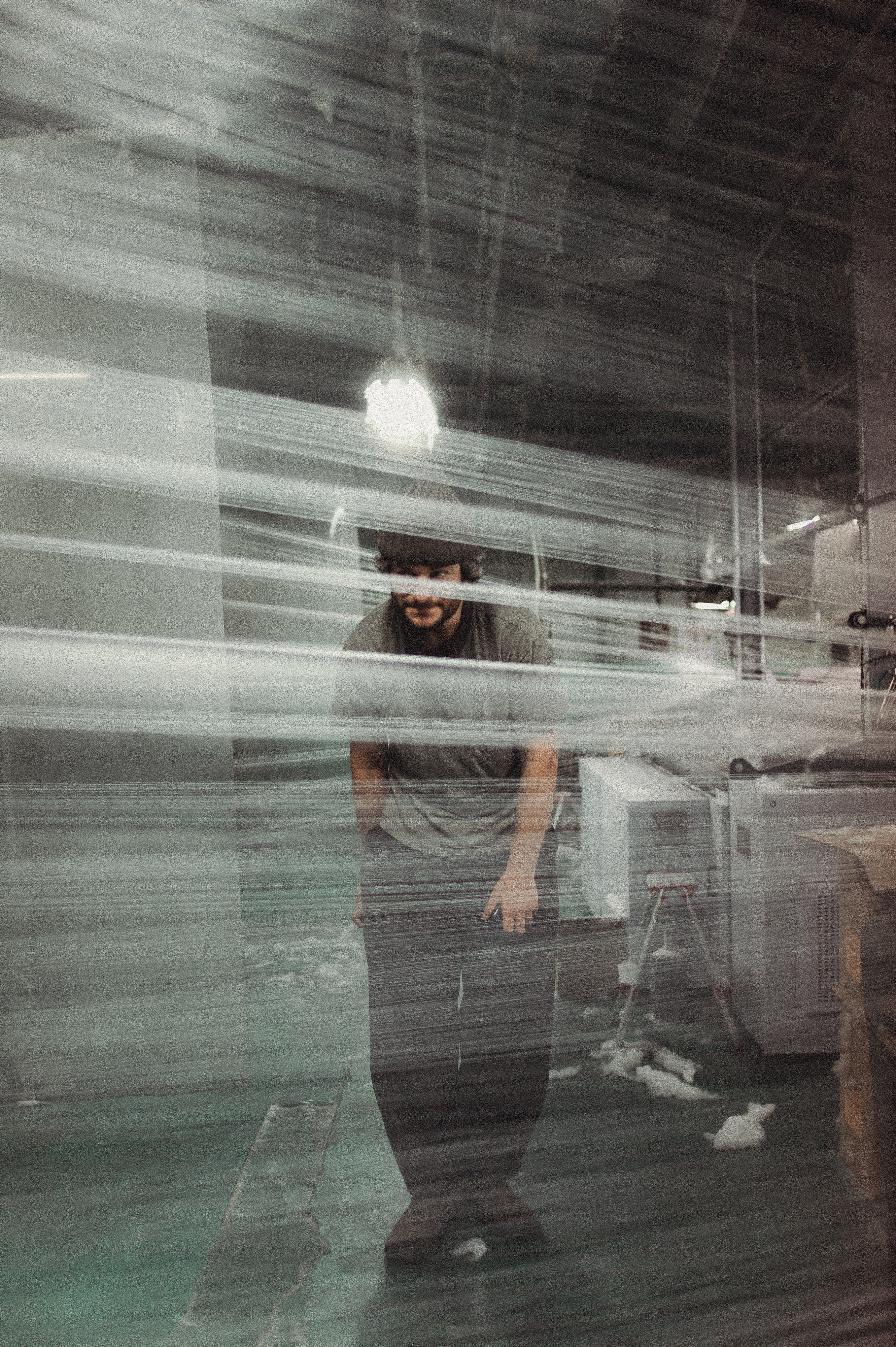 A person wearing a beanie is faintly visible behind blurred, horizontal streaks, likely plastic wrap, in an industrial setting with machinery and soft lighting.