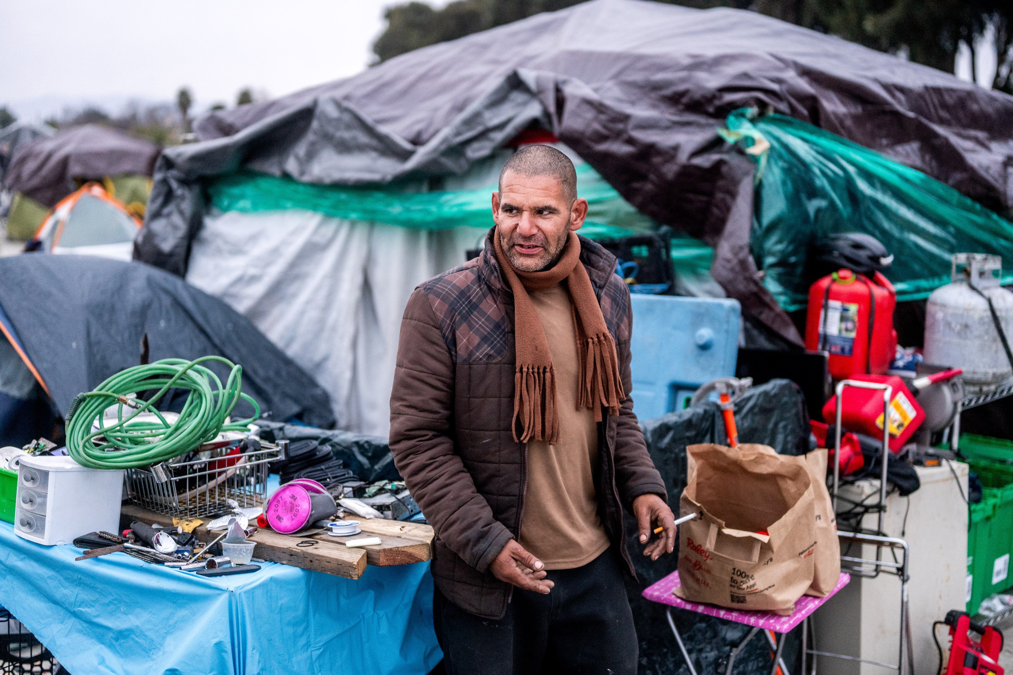 A man in a plaid jacket stands near a cluttered table with various tools and objects. Behind him are tarps covering structures and several colorful containers.