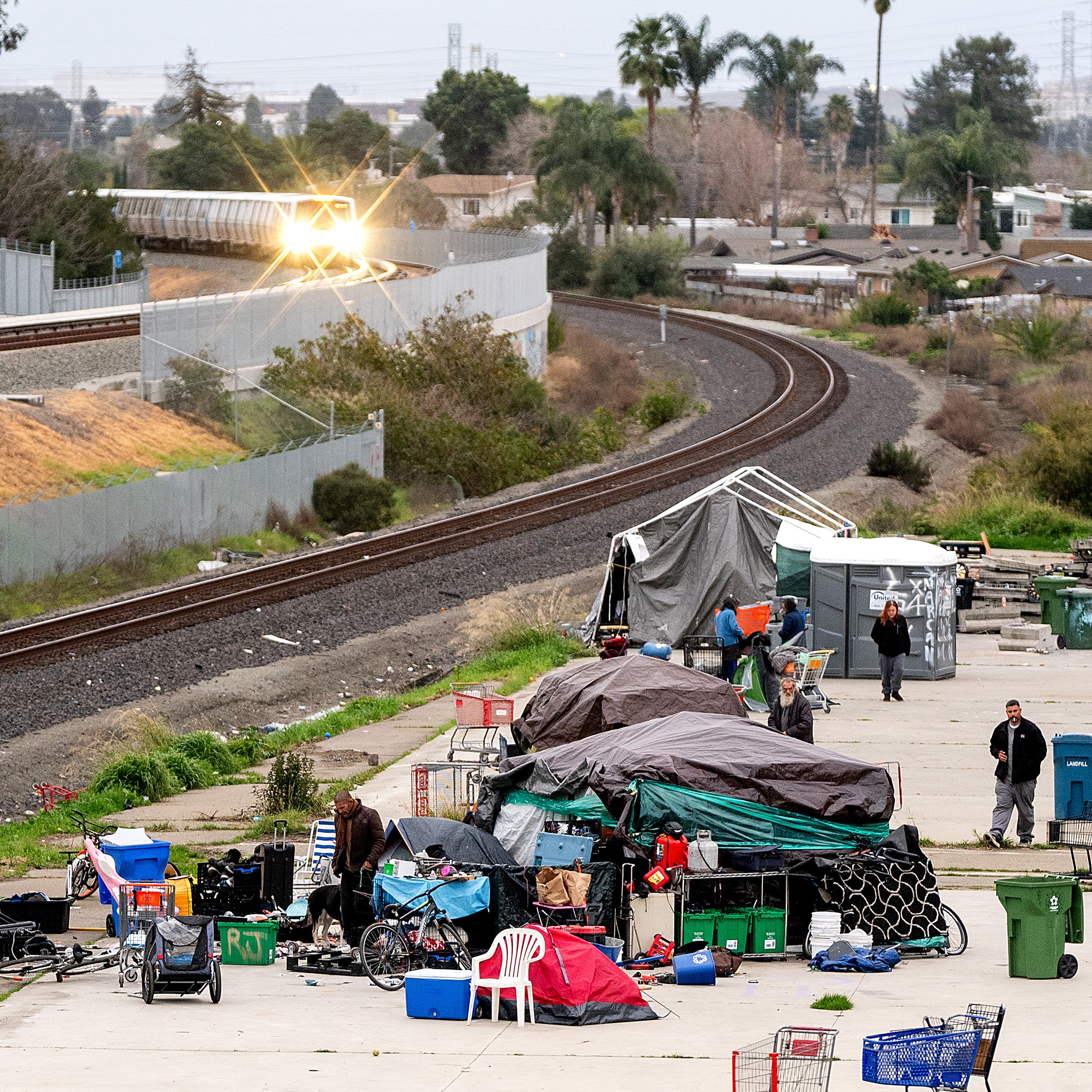 A group of tents and belongings is set up along a curved train track. People are around the makeshift area, with scattered items and several large bins.