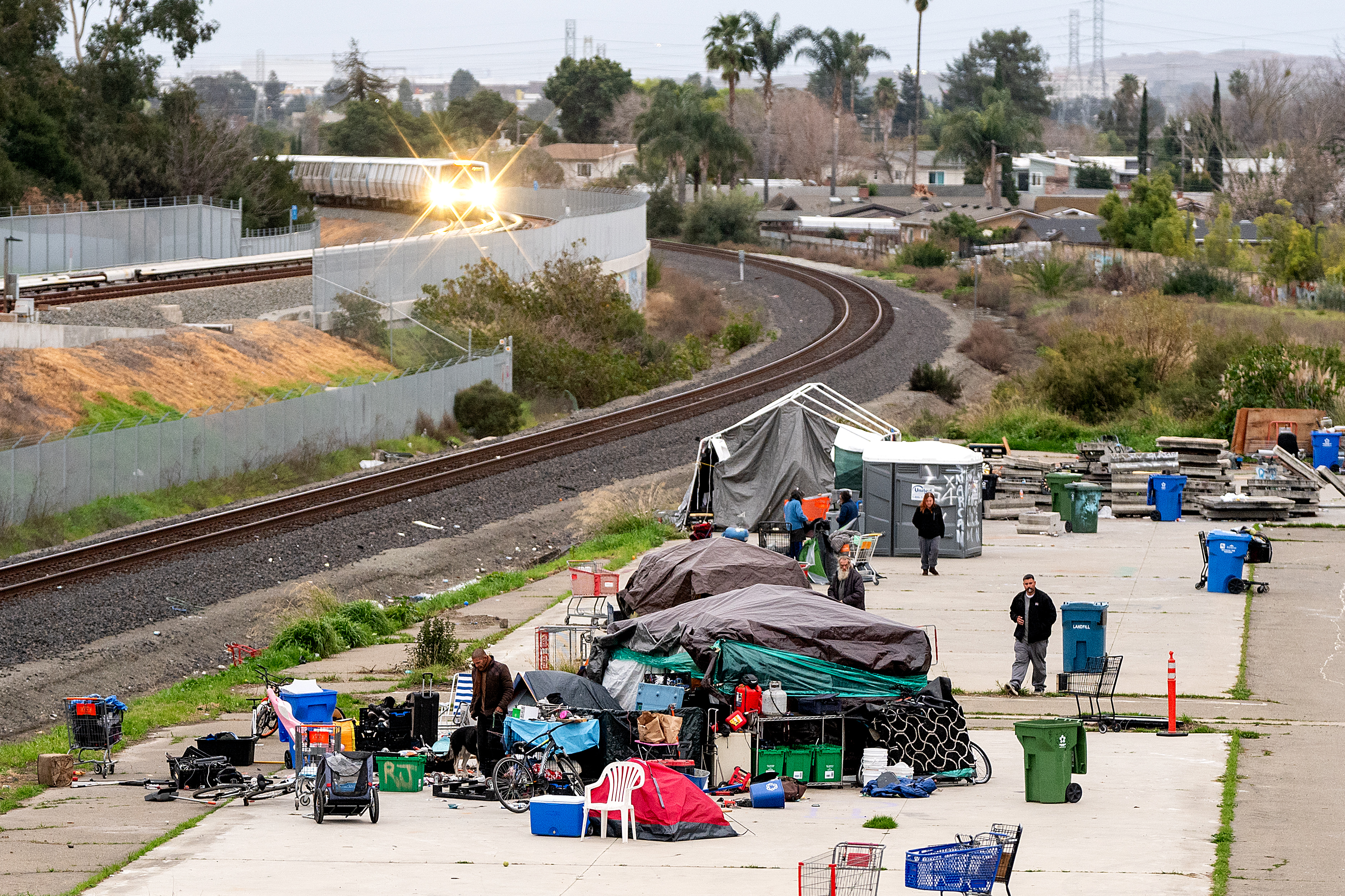 A group of tents and belongings is set up along a curved train track. People are around the makeshift area, with scattered items and several large bins.