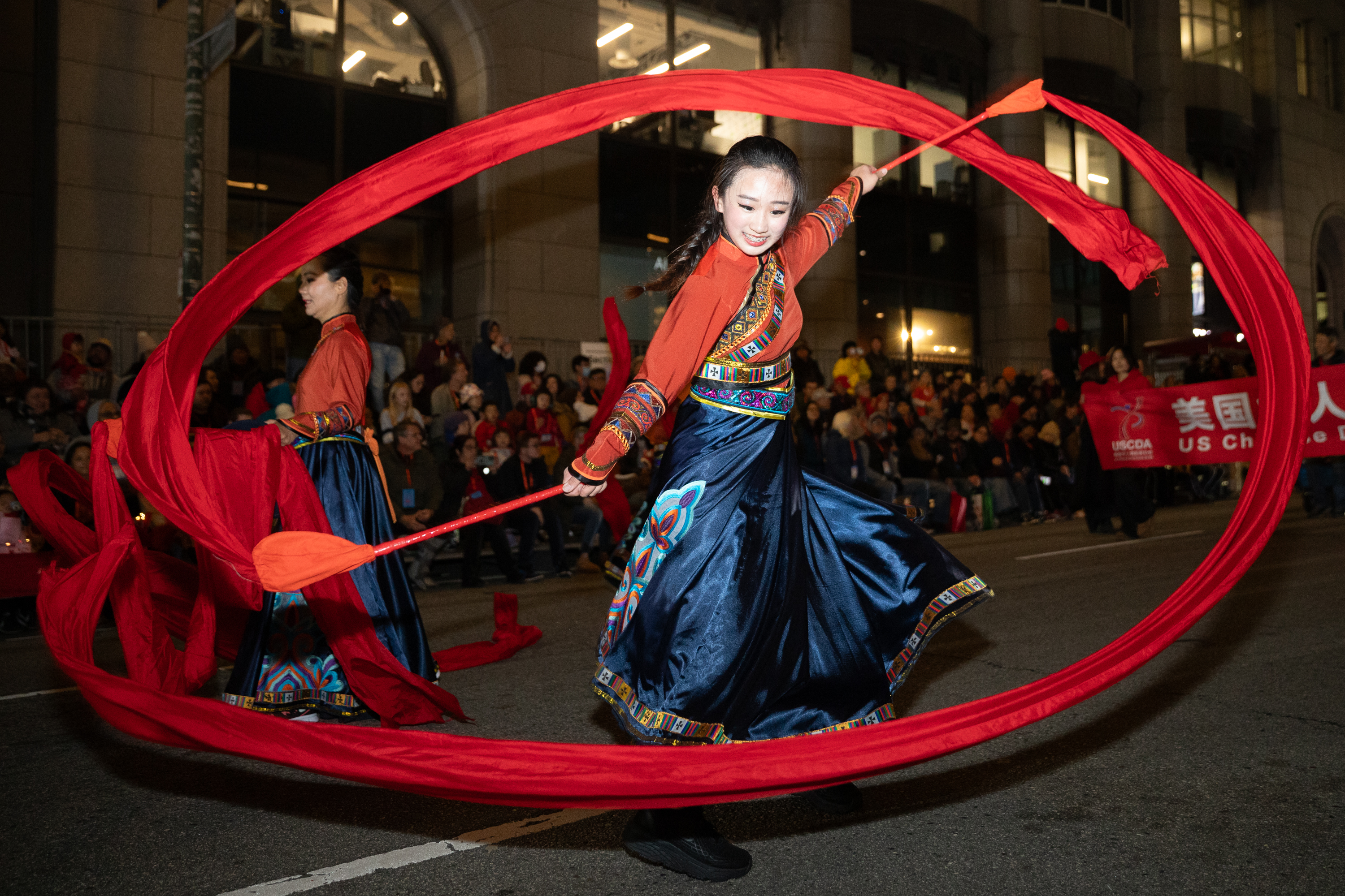 A dancer in colorful traditional attire energetically twirls a long red ribbon in a street parade, surrounded by an audience at night.