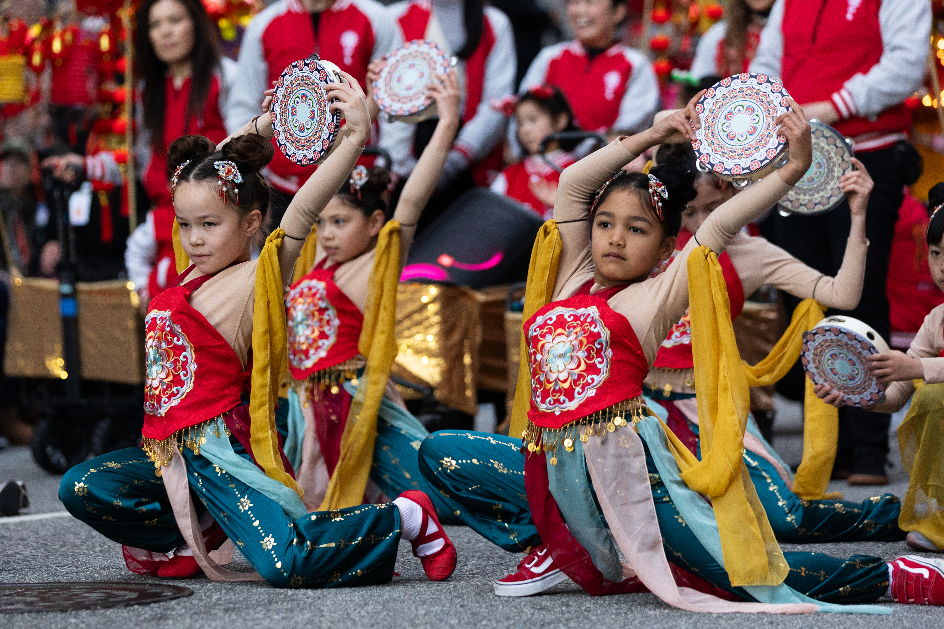 Children in colorful outfits perform a dance, holding decorated circular instruments aloft. They are kneeling on one knee with synchronized poses.