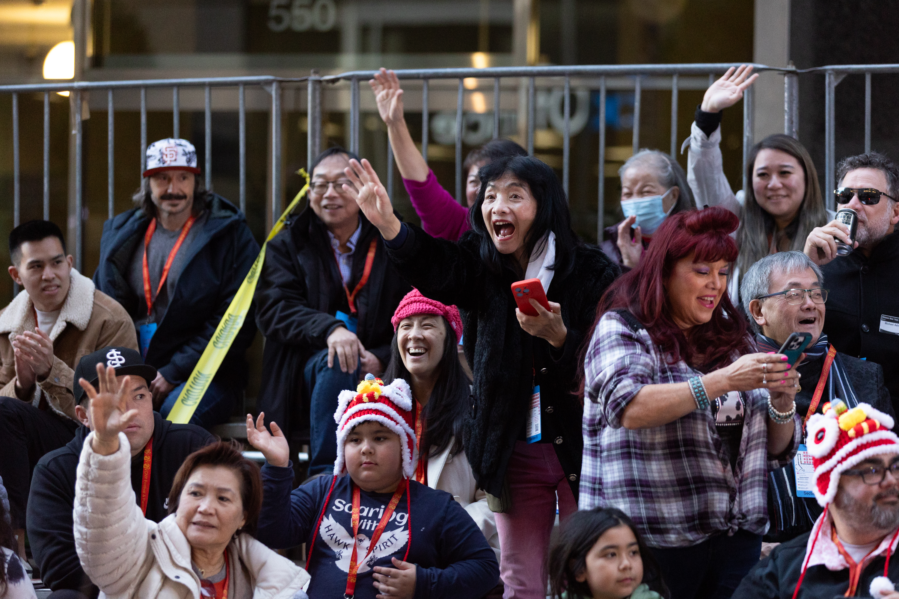 A cheerful crowd is gathered, some waving and taking photos. They appear to be outdoors, with a metal barrier behind them. One child wears a colorful hat.