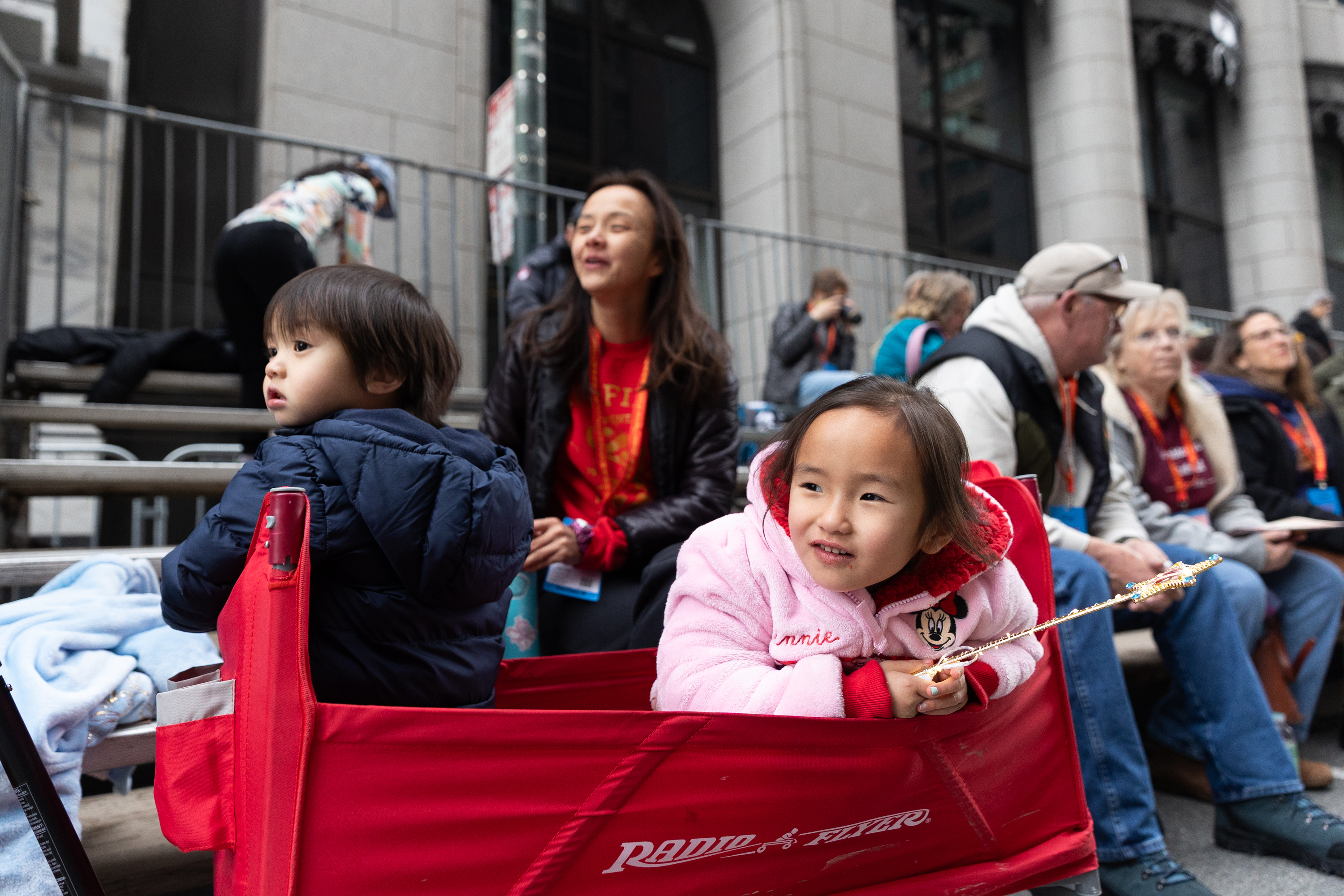 A young girl in a pink coat sits in a red wagon, smiling and holding a wand. A boy is beside her. Adults sit on bleachers in the background.