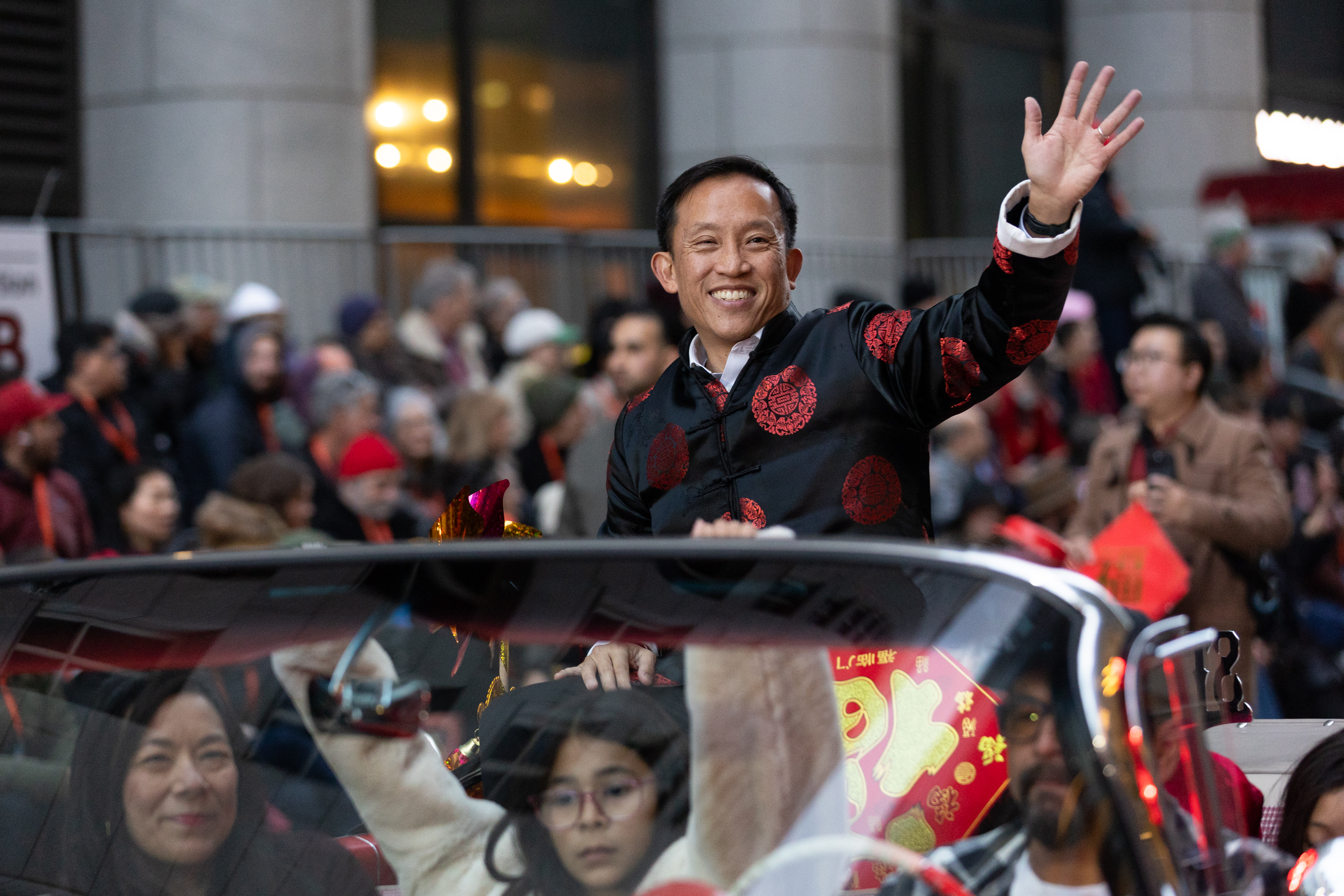 A smiling man in a black and red traditional jacket waves from a moving car in a parade. Spectators line the street, watching the event unfold.