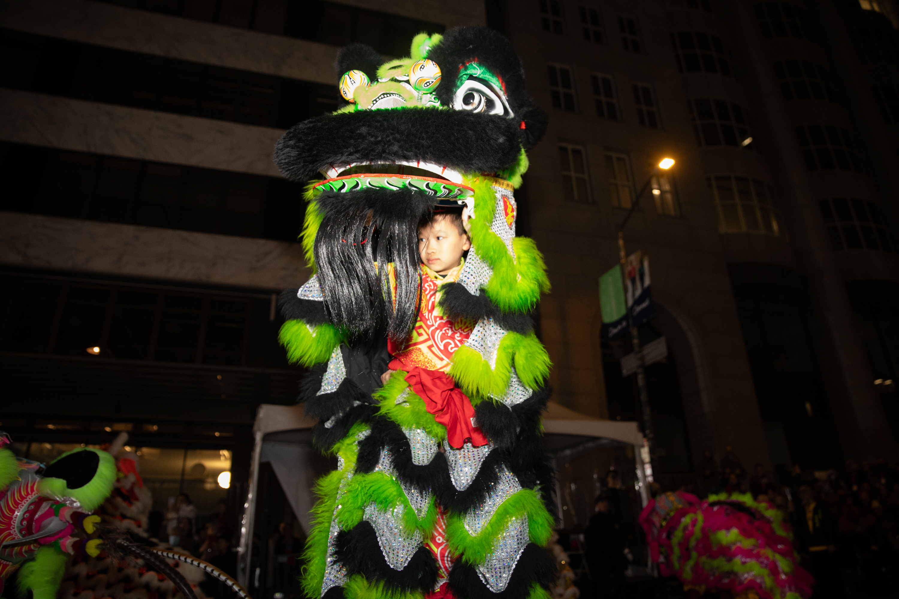 A child peeks out from a colorful, elaborate dragon costume, featuring green and black fur, during a nighttime performance on a city street.