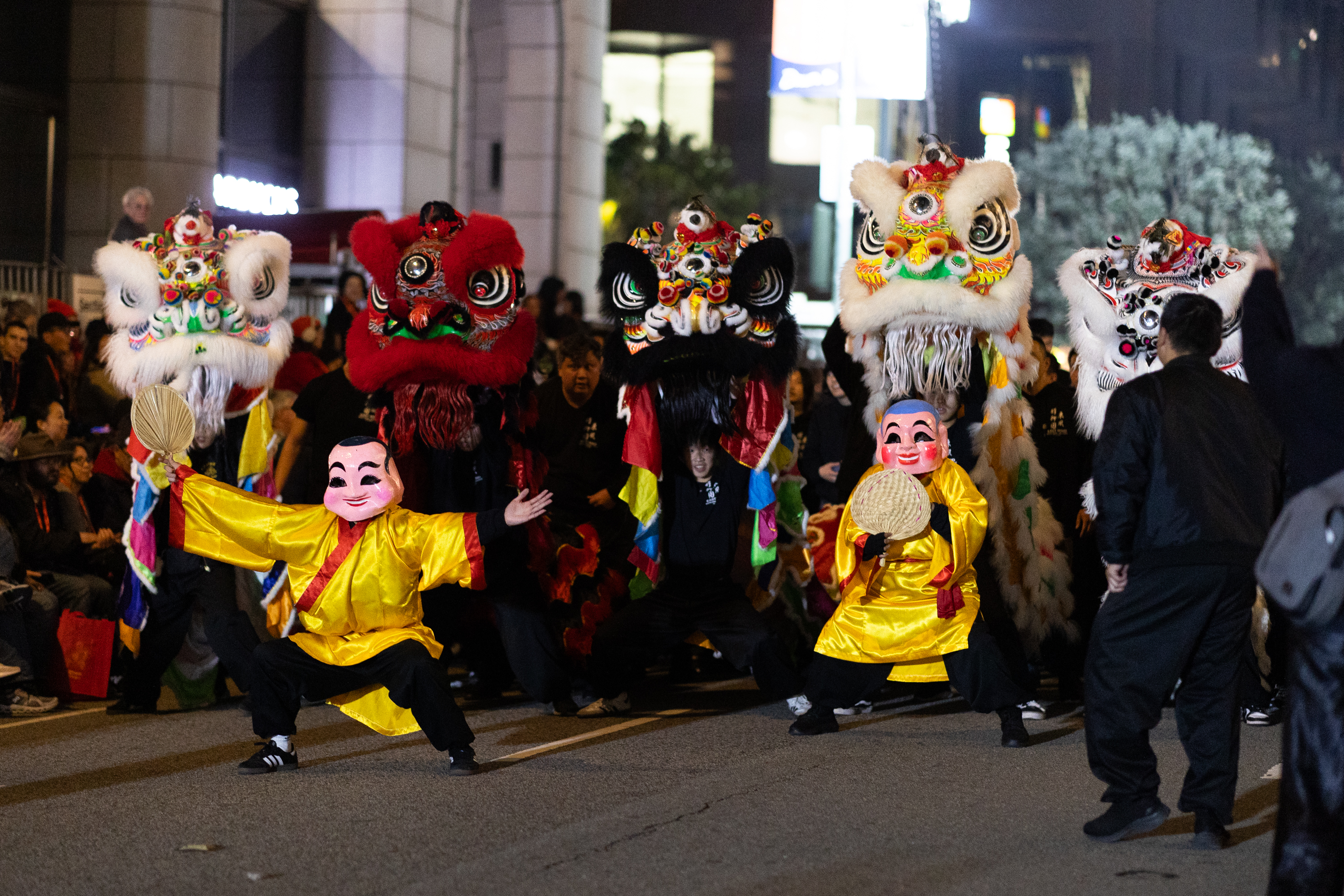 Performers in colorful lion costumes dance energetically, accompanied by people wearing vibrant masks and traditional attire, in a lively street festival.