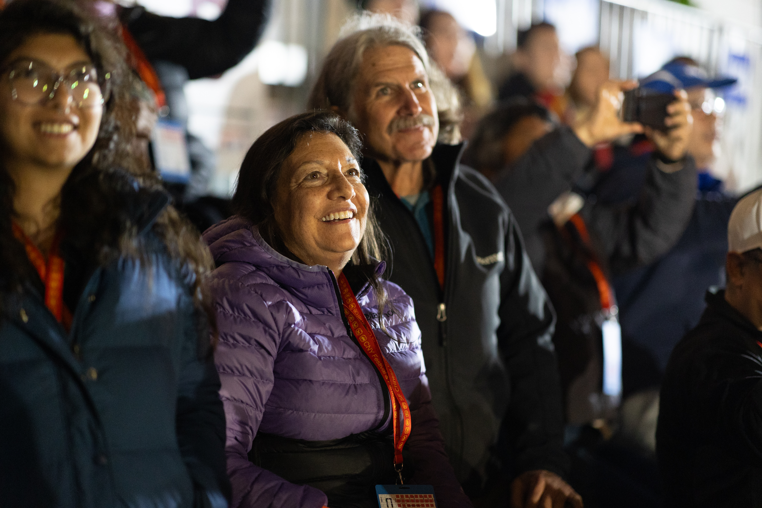 A group of people is gathered, looking upward with smiles. The woman in the front is wearing a purple jacket. The background is blurred, with someone taking a photo.