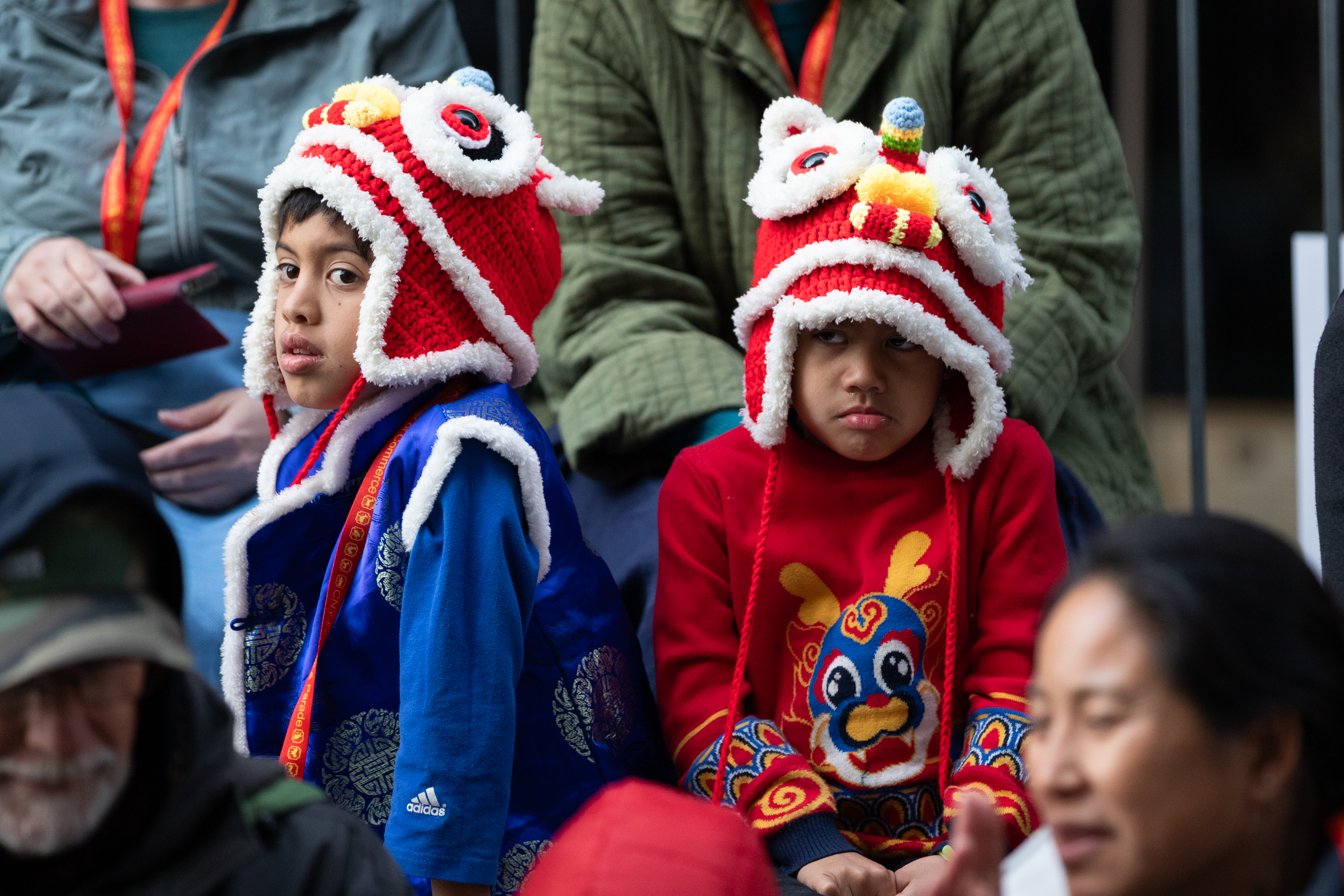 Two children are seated, wearing vibrant lion dance hats. One child wears blue with a festive design, while the other sports a patterned red outfit.