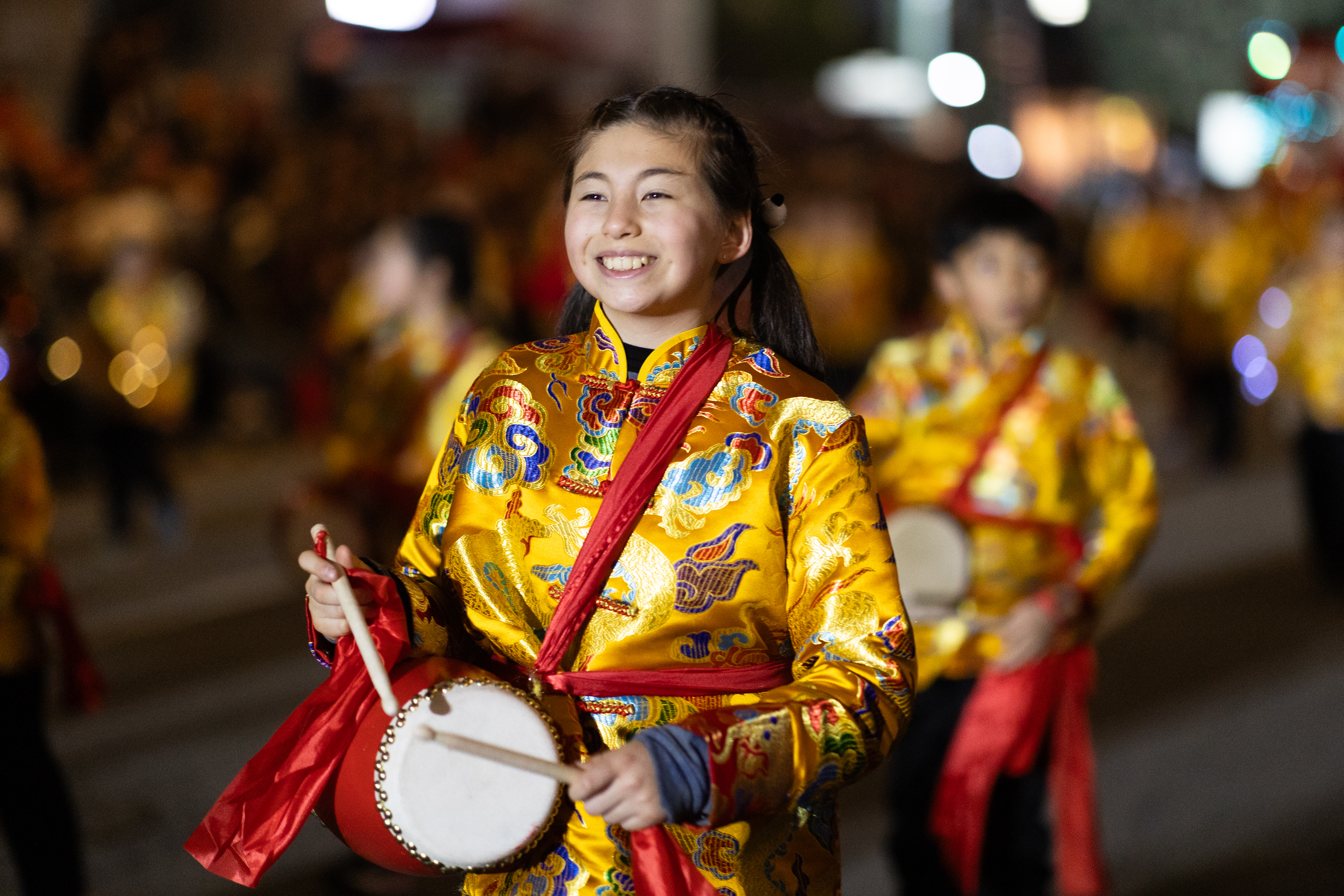 A young person in a bright, embroidered yellow outfit plays a handheld drum at a night parade, smiling amidst a group of similarly attired participants.