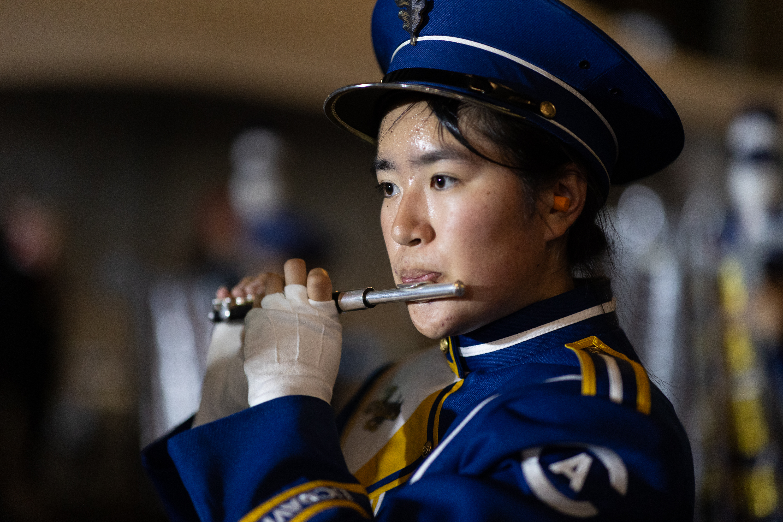 A person in a blue marching band uniform and hat plays a flute, with a focused expression, while wearing gloves. The background is blurred.