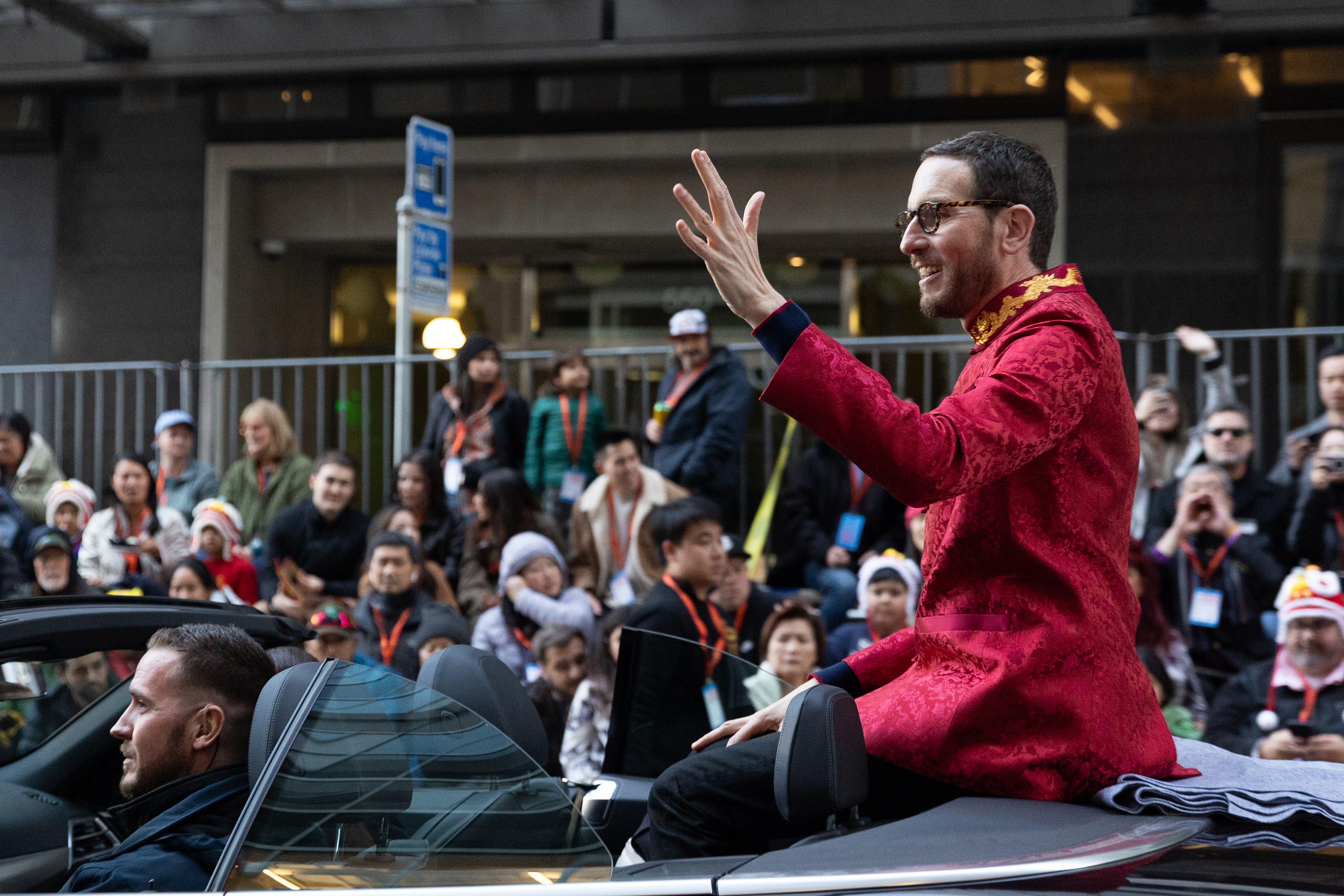 A person in a red ornate outfit waves while sitting on the back of a convertible during a parade. People in the background watch and take photos.