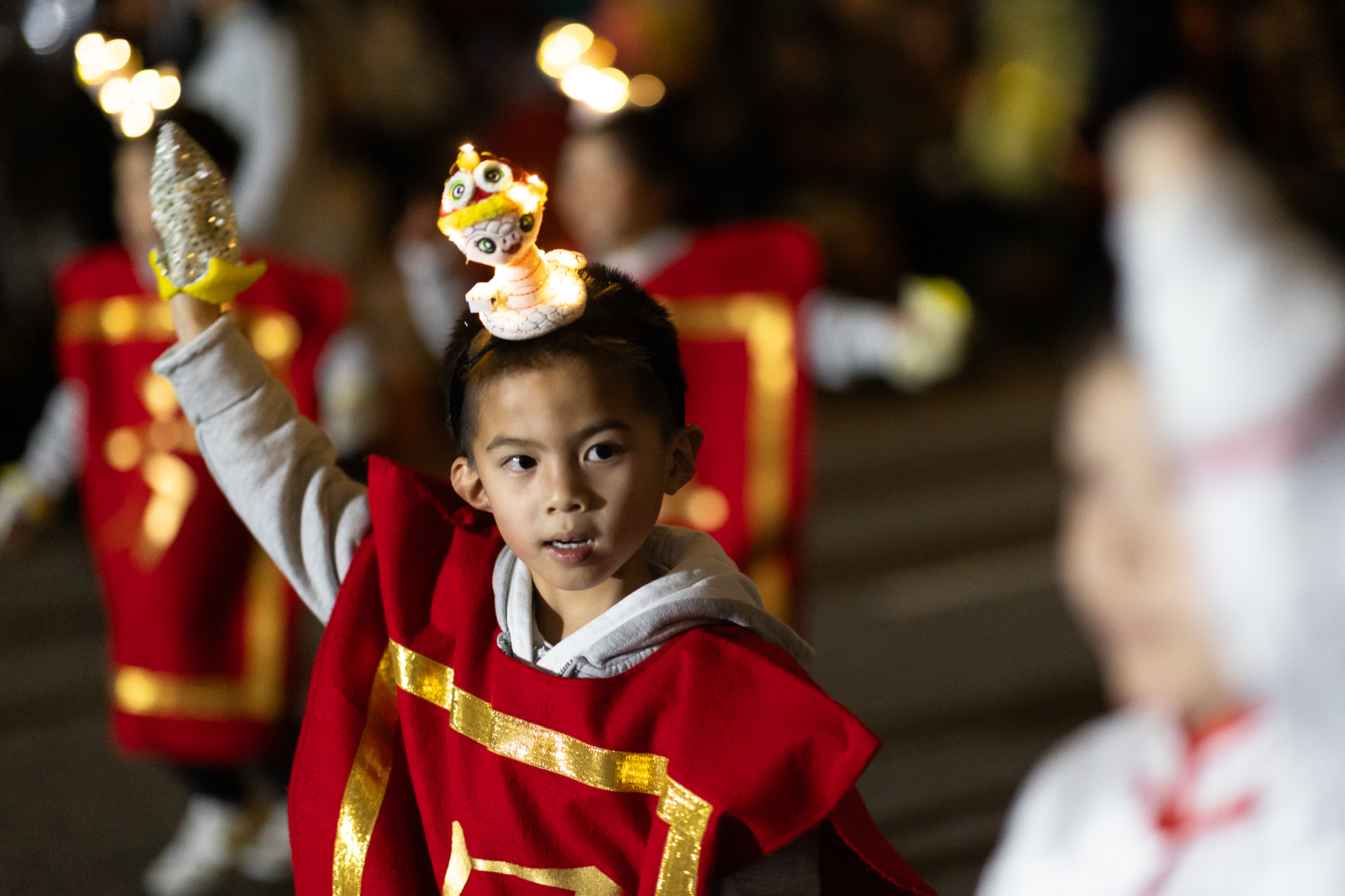 A child wearing a red costume with gold trim raises one arm. A small lit headpiece resembling a dragon is on their head, with a blurred background suggesting a parade.