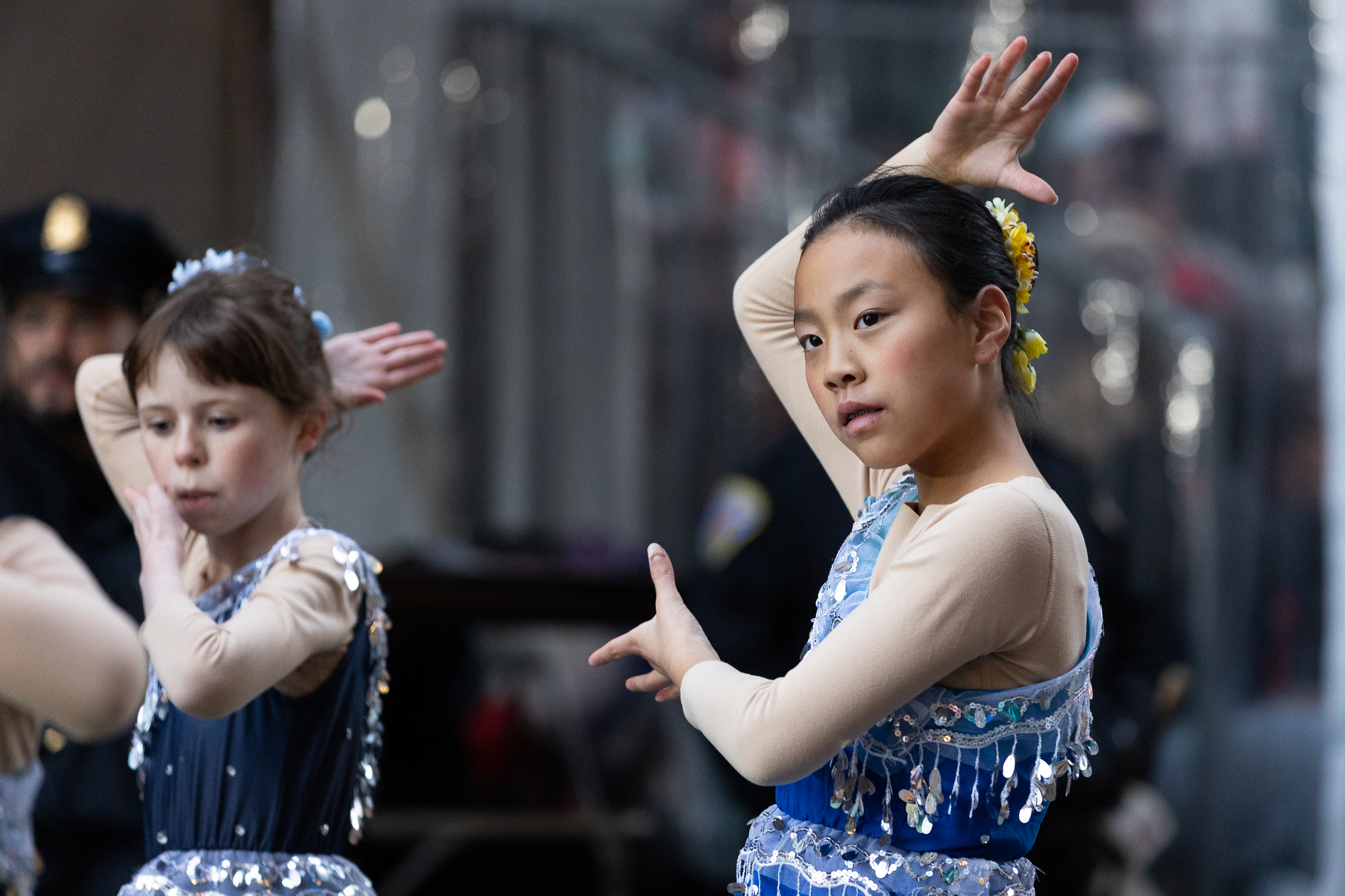 Two young dancers perform, wearing blue sequin costumes and beige long-sleeve tops. One has a flower in her hair, and both have their arms gracefully posed.