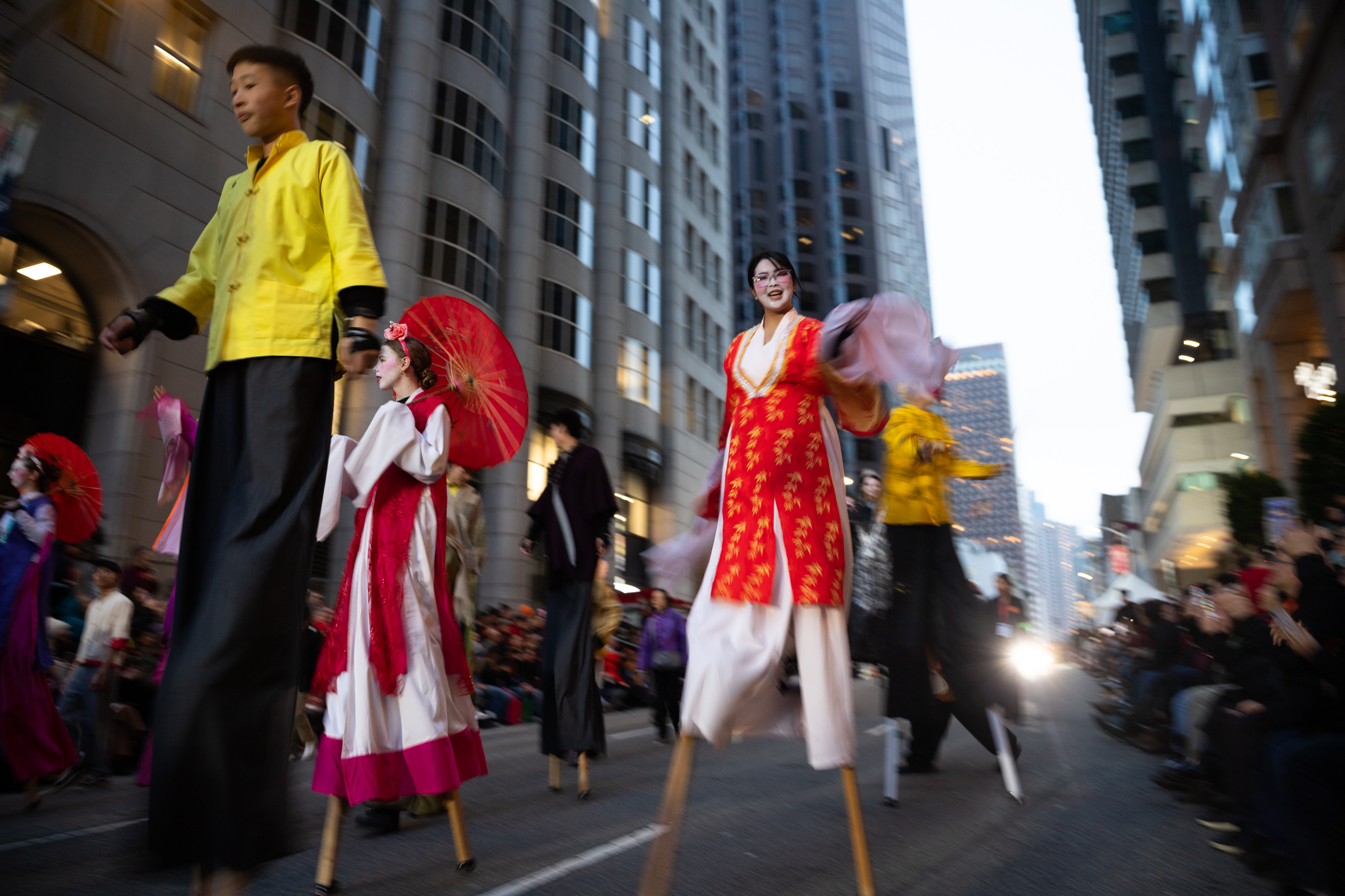 People on stilts in colorful traditional costumes parade down a city street. Tall buildings and a crowd of spectators are visible in the background.