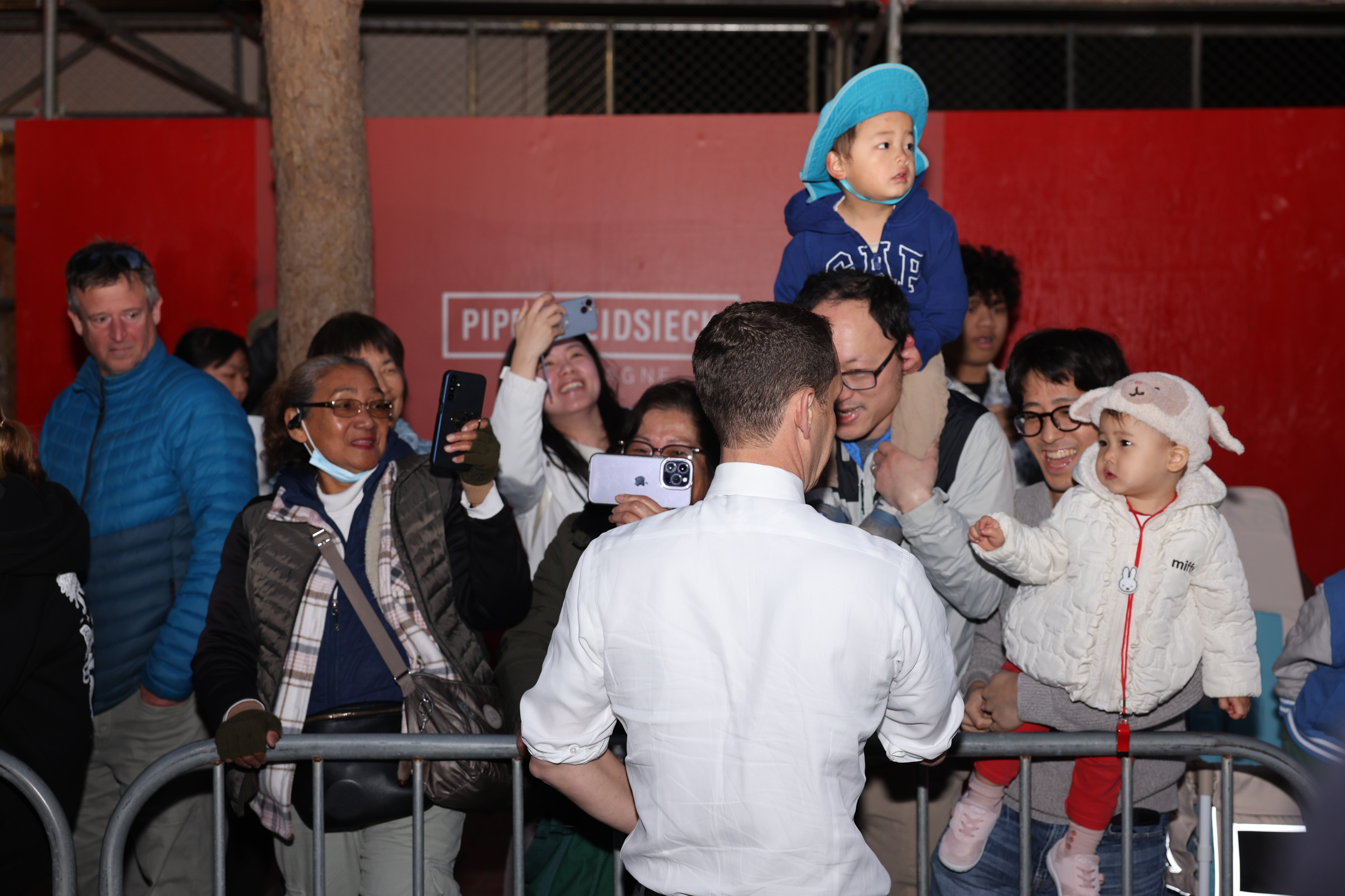 A crowd joyfully snaps photos while a man in a white shirt interacts with them. Two children are being held, one on a shoulder, the other in arms.