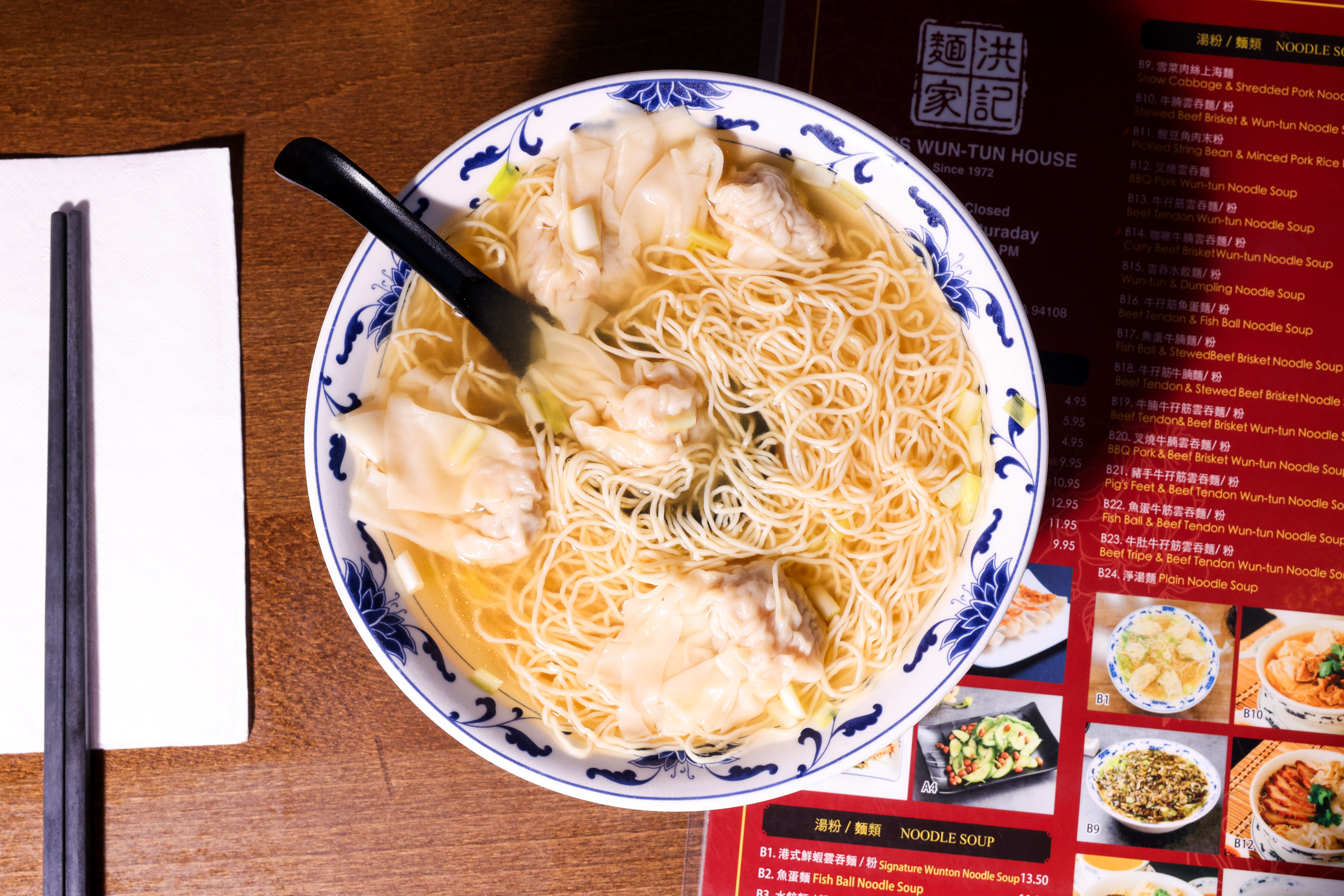 A bowl of noodle soup with dumplings sits on a wooden table, accompanied by chopsticks and a menu nearby. A napkin and a black spoon are also visible.