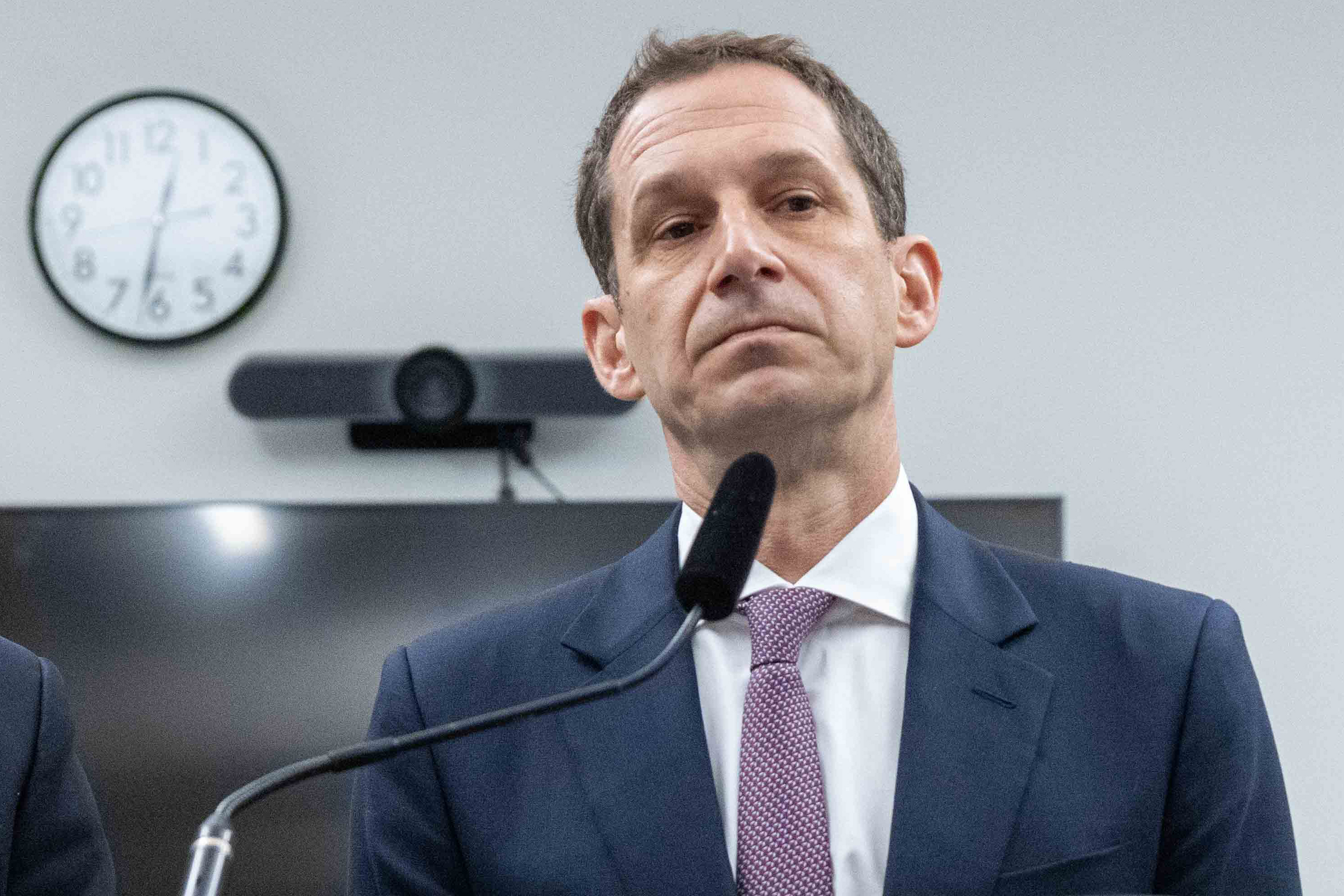 A man in a suit stands in front of a microphone, with a wall clock and a screen behind him. His expression is serious.