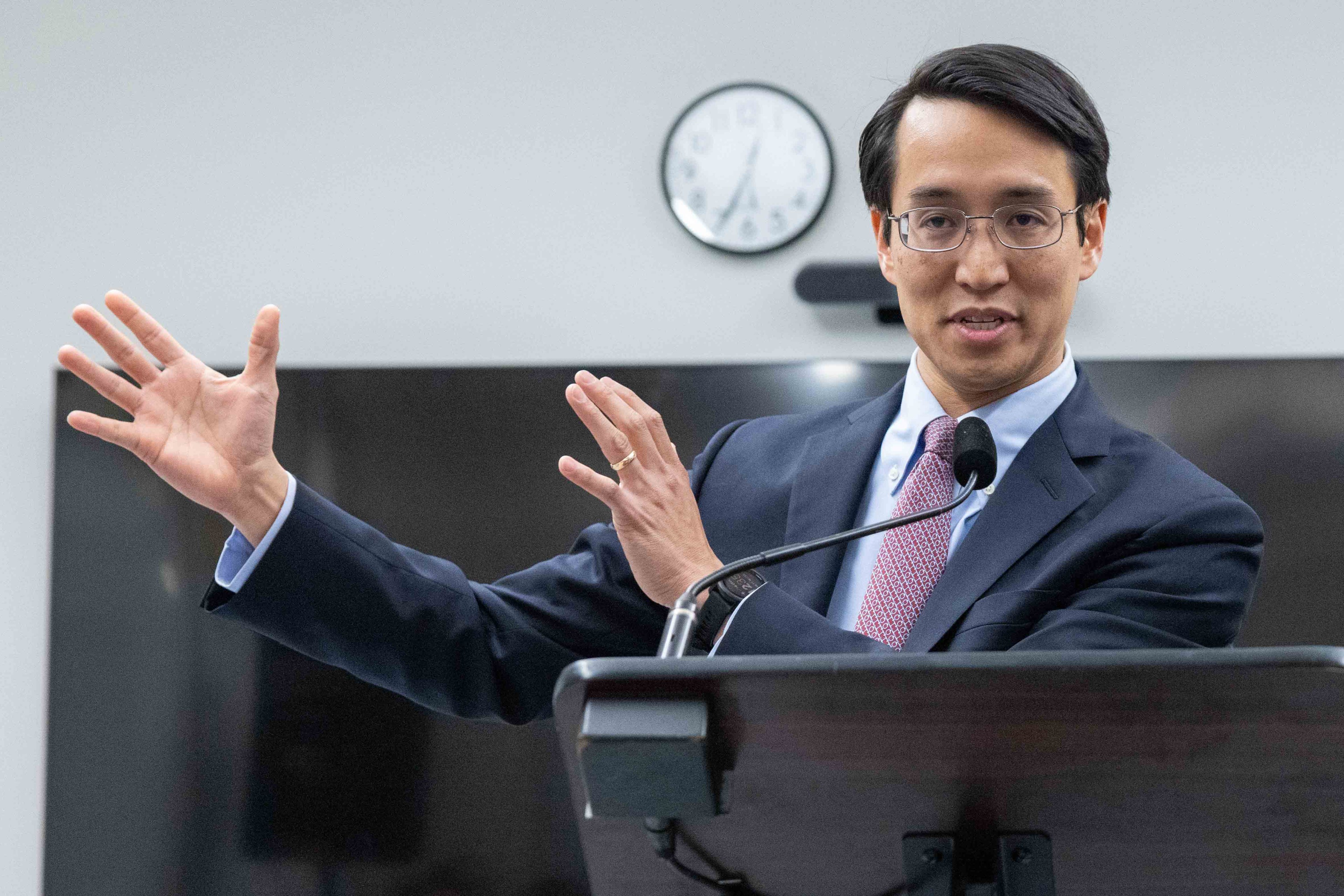 A man in a suit speaks at a podium. He gestures with his hands, and there's a microphone in front of him. A clock and a TV screen are visible in the background.